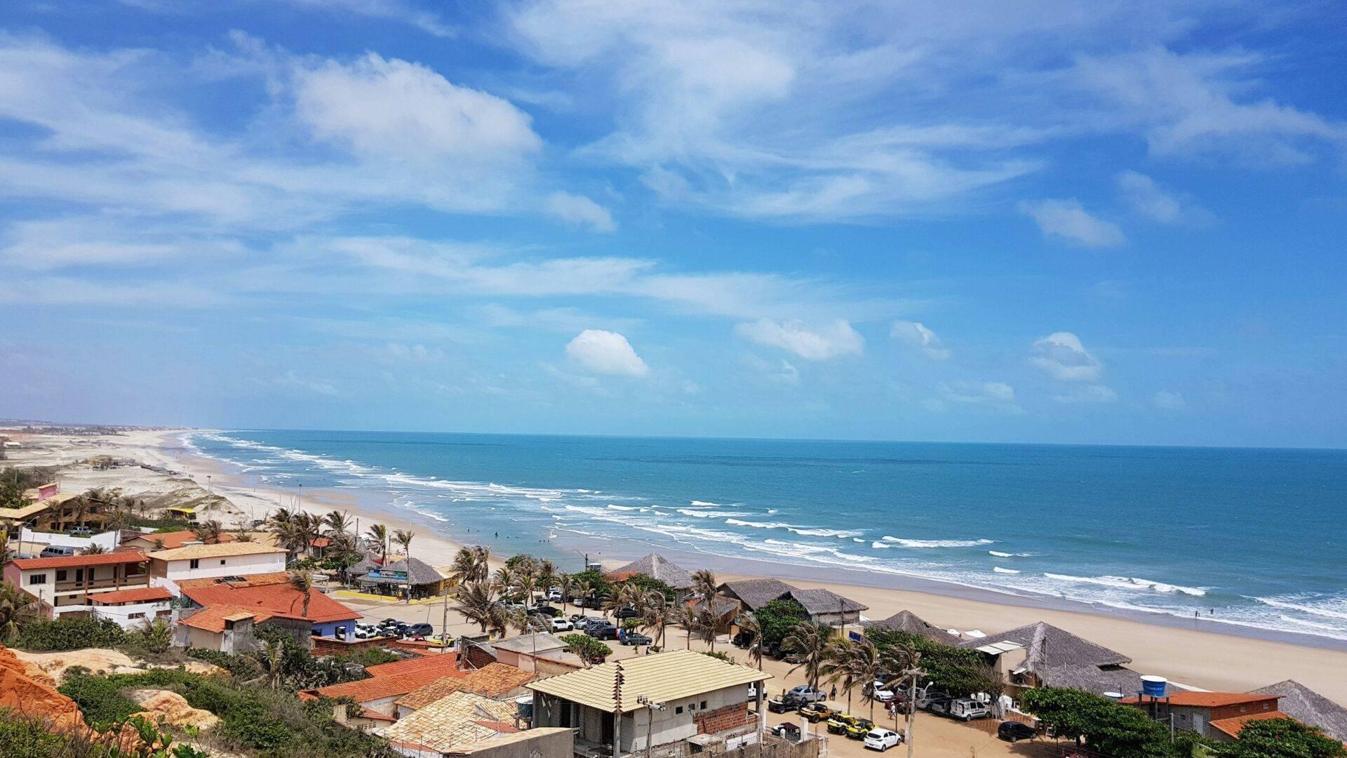 Aerial view of a white sand beach with waves, colourful houses with orange roofs and palm trees