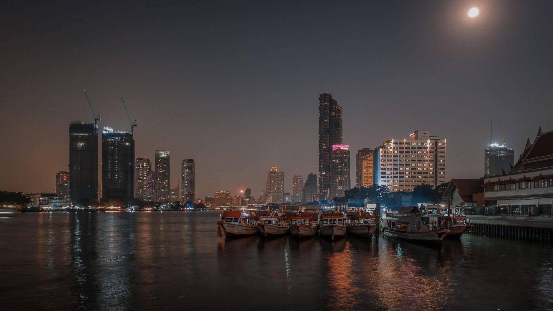 Skyline urbain illuminé la nuit avec bateaux traditionnels rouges amarrés devant une architecture moderne au bord d’un fleuve