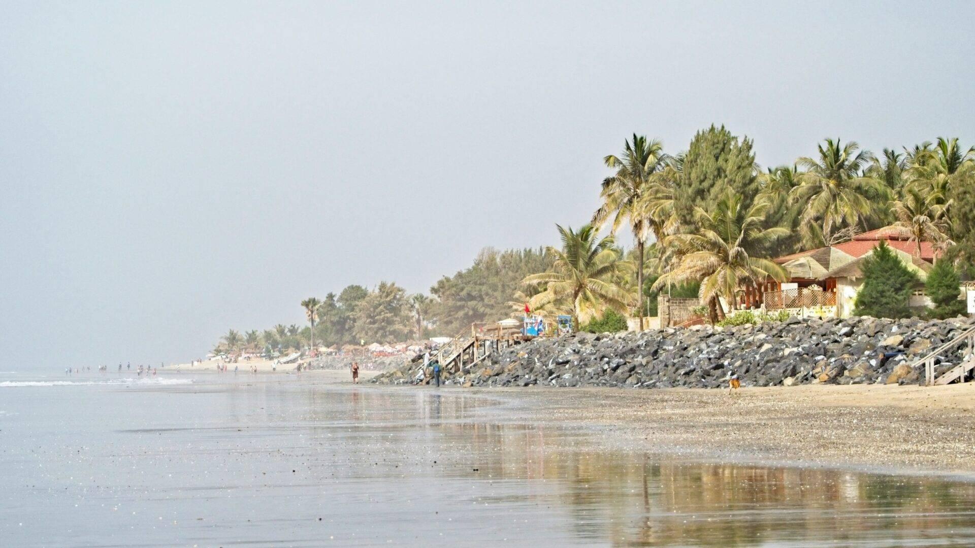 Una spiaggia costiera con palme, edifici colorati e detriti rocciosi, immersa nella luce del tardo pomeriggio
