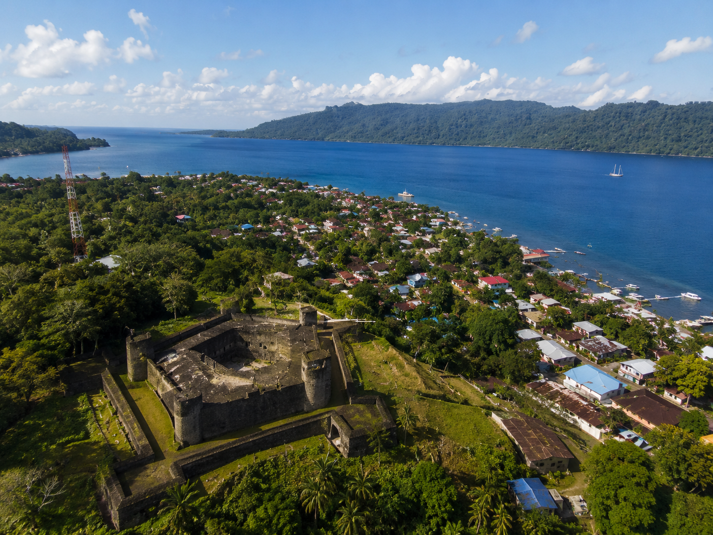 Forteresse coloniale surplombant une baie turquoise avec village côtier et forêt tropicale dense