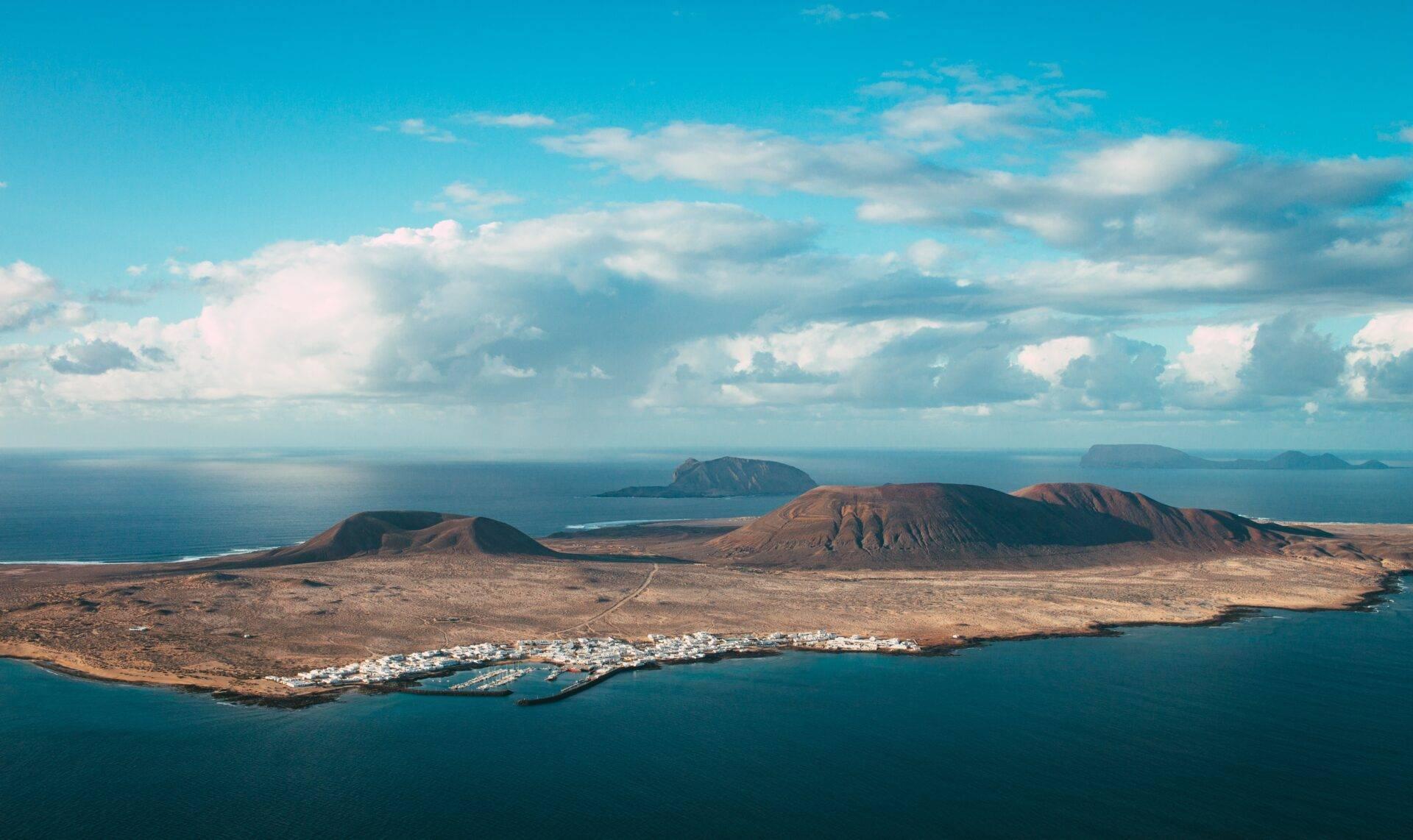 Vue aérienne d’une île volcanique aride avec petite ville côtière blanche, mer turquoise et îles lointaines sous ciel dégagé