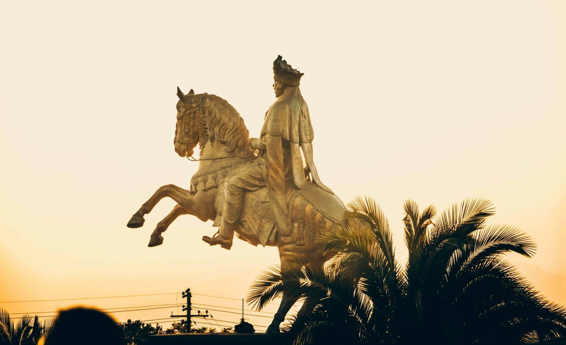 Goldene Reiterstatue eines Königs in königlichem Gewand bei Sonnenuntergang, umgeben von Palmen