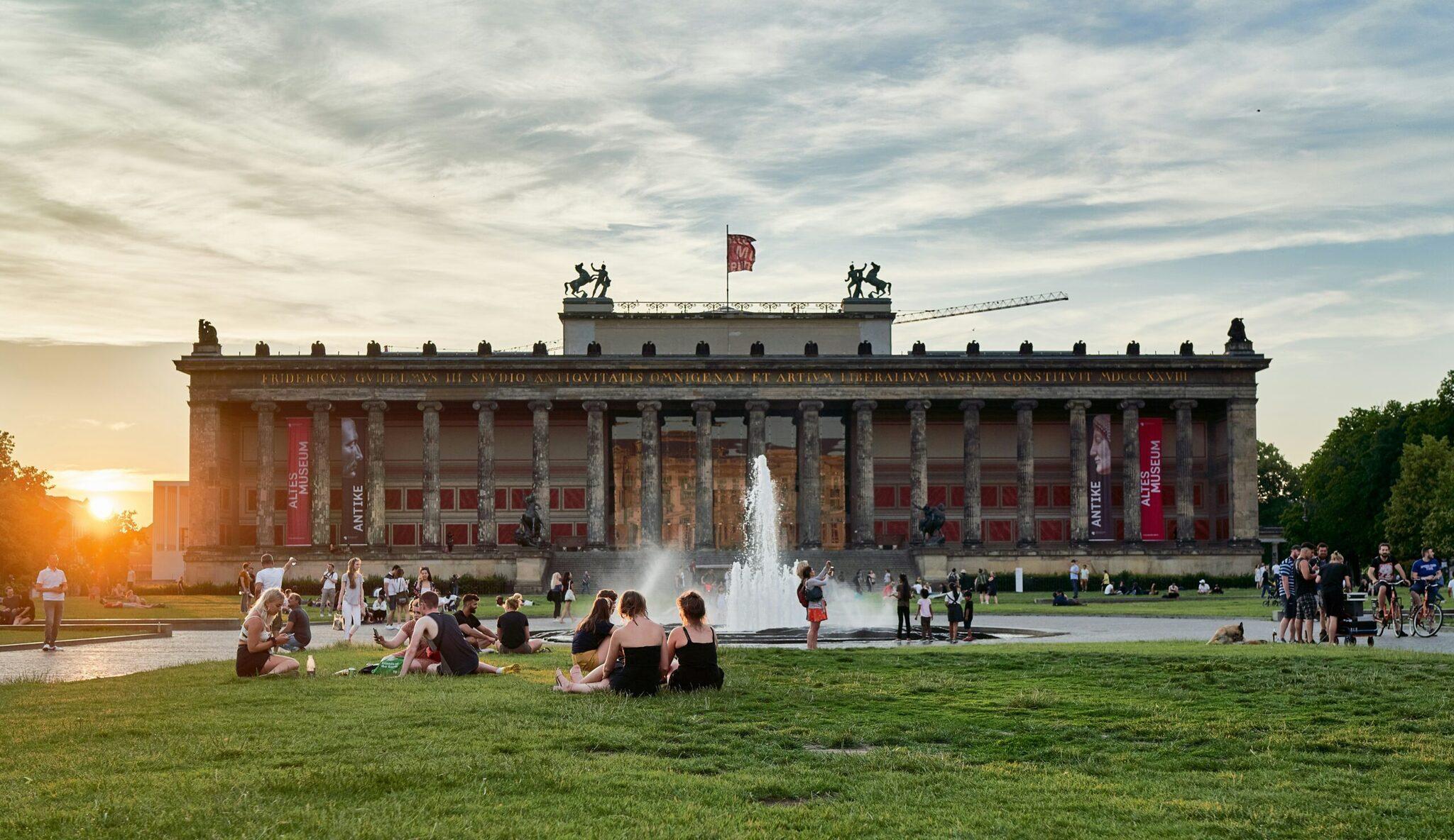 A neoclassical museum with imposing columns and a fountain, surrounded by visitors relaxing on the lawn at sunset