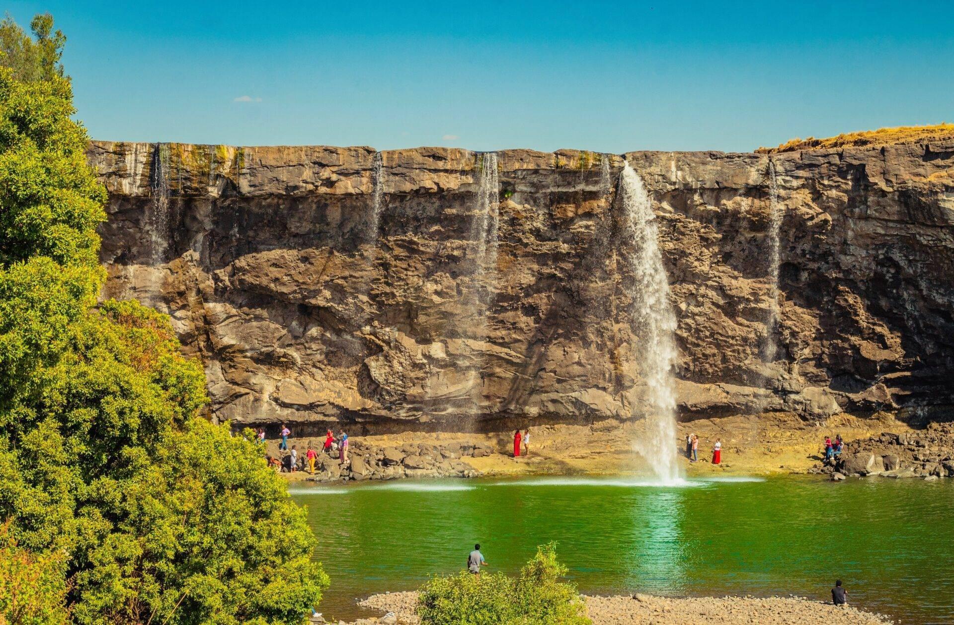 Ein spektakulärer Wasserfall stürzt von einer Felswand in einen türkisfarbenen See, umgeben von Besuchern und üppiger Vegetation