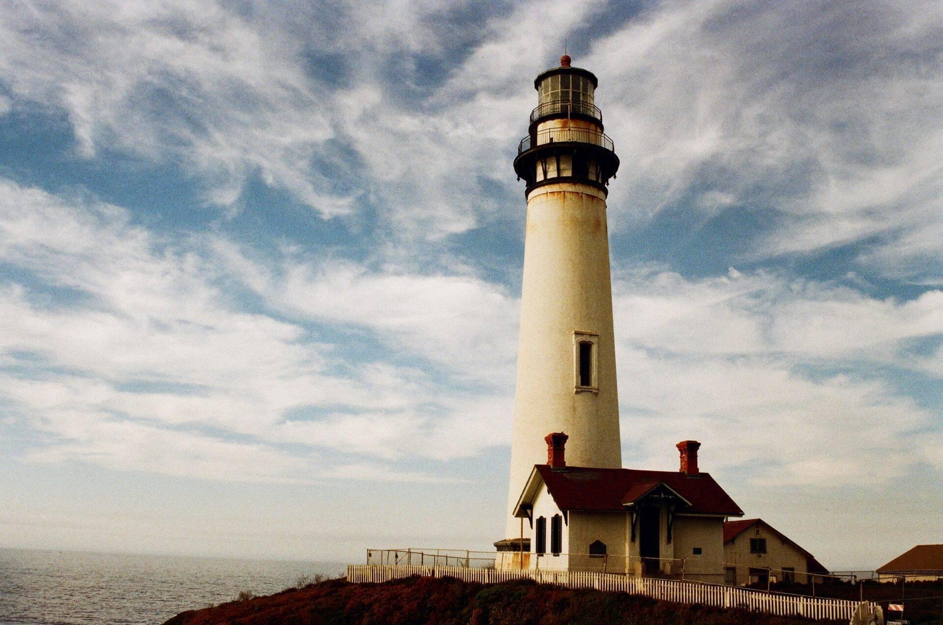 Un phare blanc côtier avec sa lanterne et ses bâtiments d’habitation aux toits rouges sous un ciel nuageux