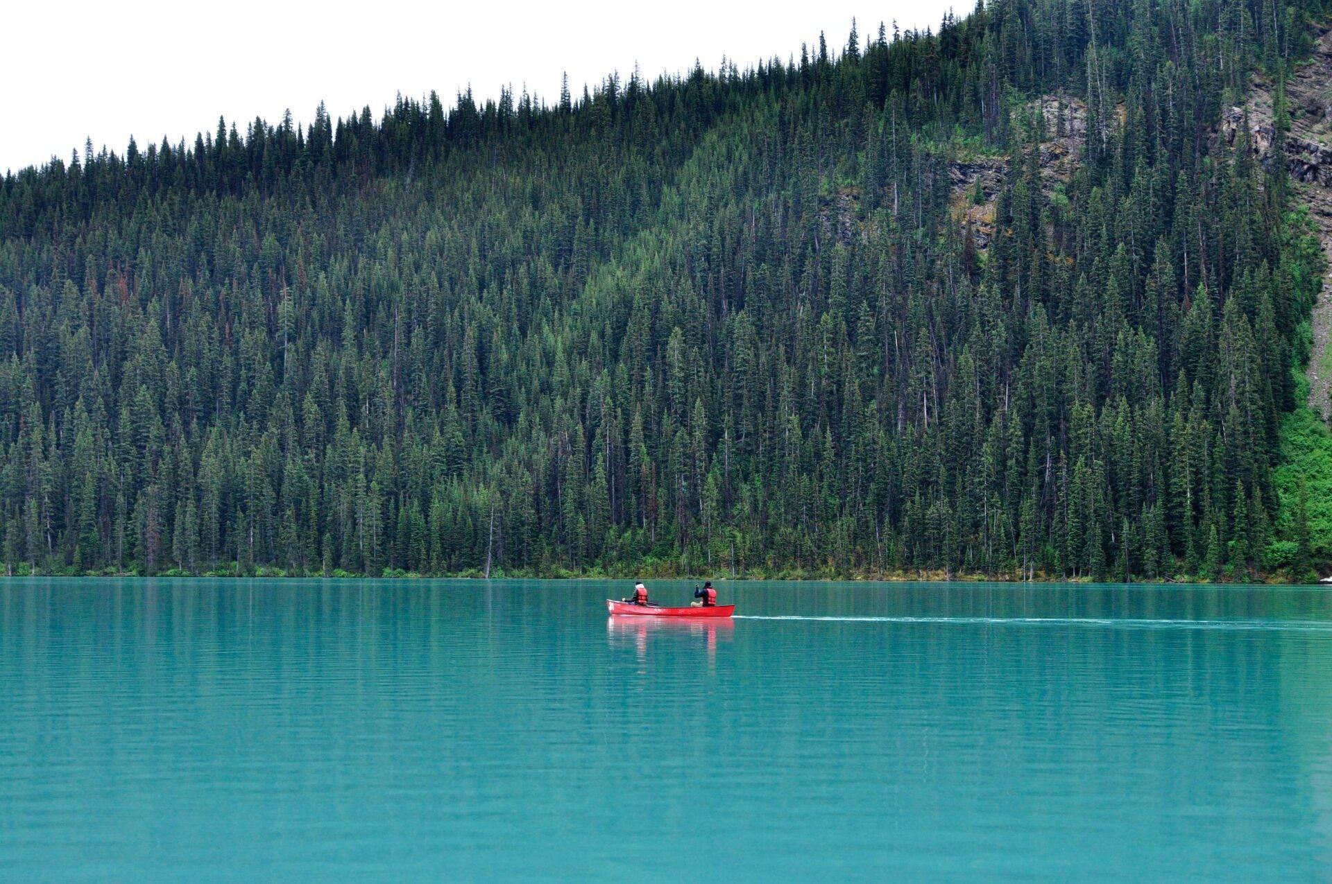 Deux personnes en canot rouge naviguent sur un lac turquoise entouré d’une forêt de conifères dense et montagneuse