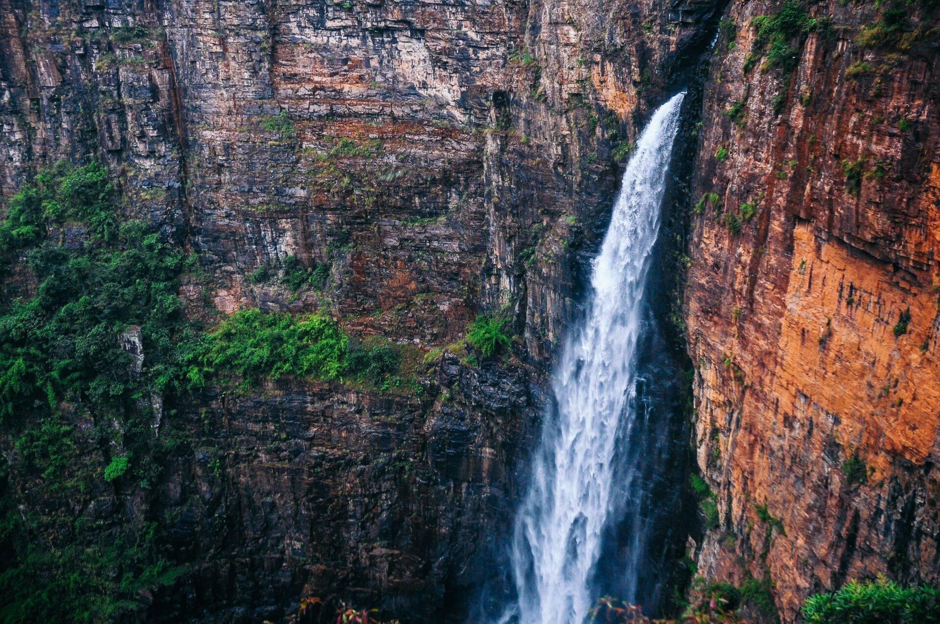 Ein weißer Wasserfall stürzt eine rötliche Felswand hinab, die von üppiger grüner Vegetation bewachsen ist