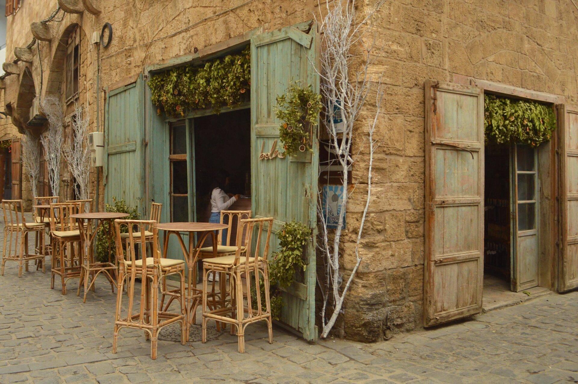 Terrasse de café avec chaises en rotin devant une façade en pierre dorée et portes vertes patinées