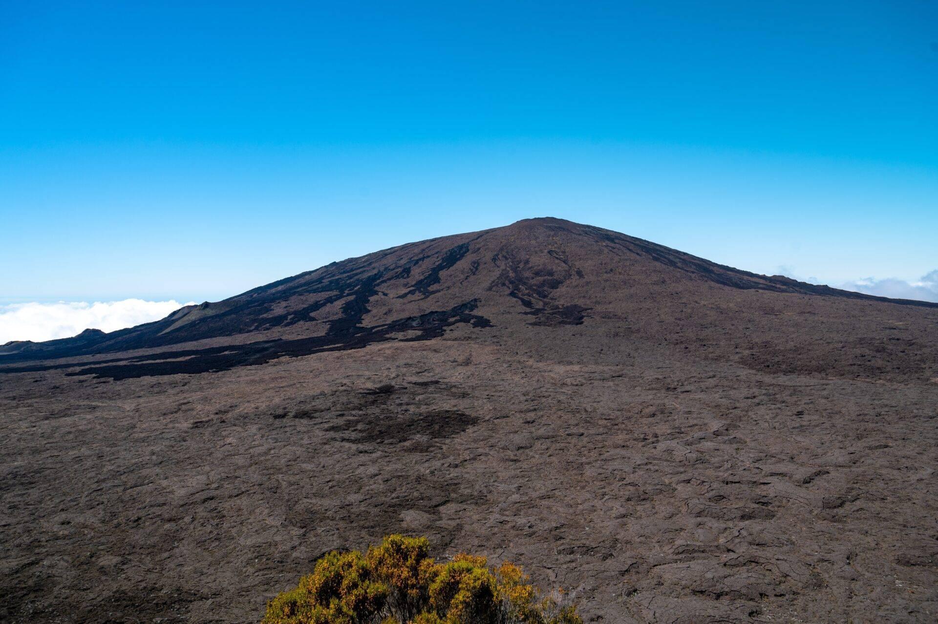 Vulkankegel mit dunklen, felsigen Hängen unter klarem blauem Himmel und gelblicher Vegetation im Vordergrund