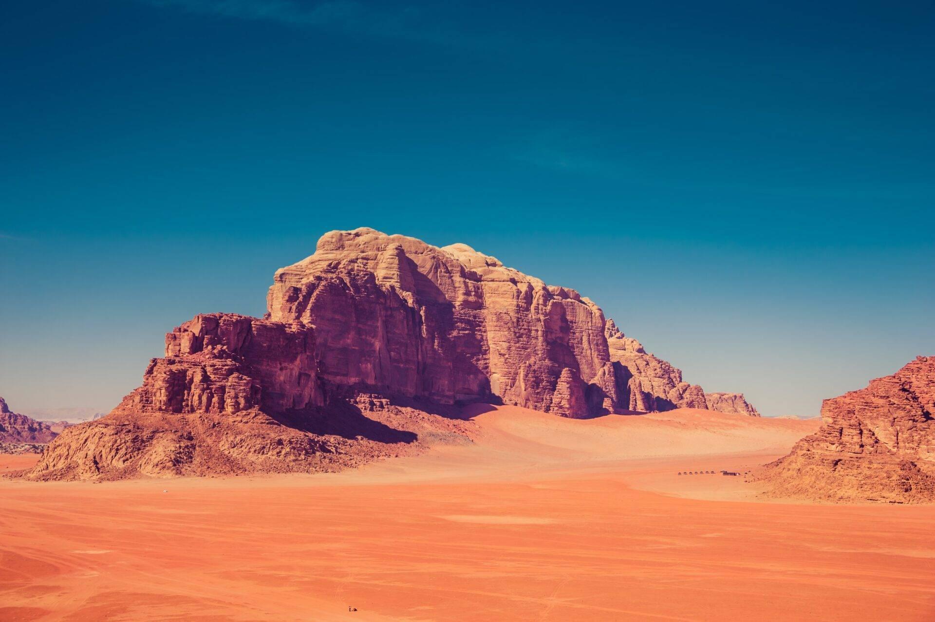 Un massiccio roccioso rosso ocra si erge in un deserto di sabbia rossastra sotto un cielo azzurro terso
