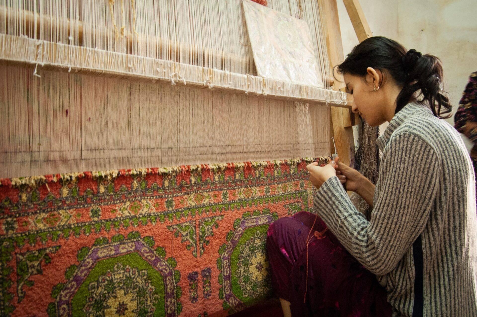 Une artisane observe attentivement la fabrication d’un tapis traditionnel coloré sur un métier à tisser manuel