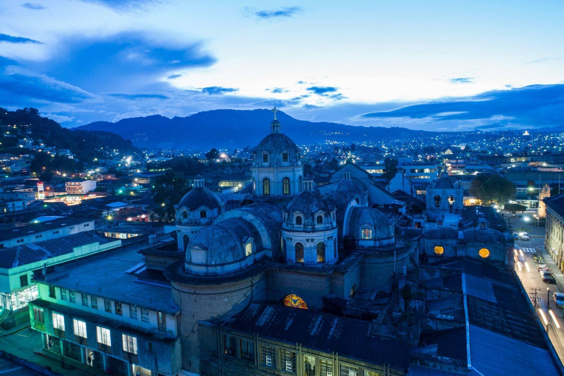 Vue aérienne d’une cathédrale coloniale aux dômes bleus illuminée au crépuscule, entourée de montagnes et de maisons
