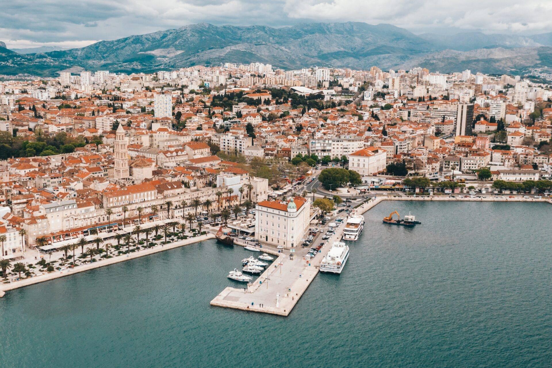 Luftaufnahme einer mediterranen Küstenstadt mit Hafen, Terrakottadächern und begrünten Bergen im Hintergrund