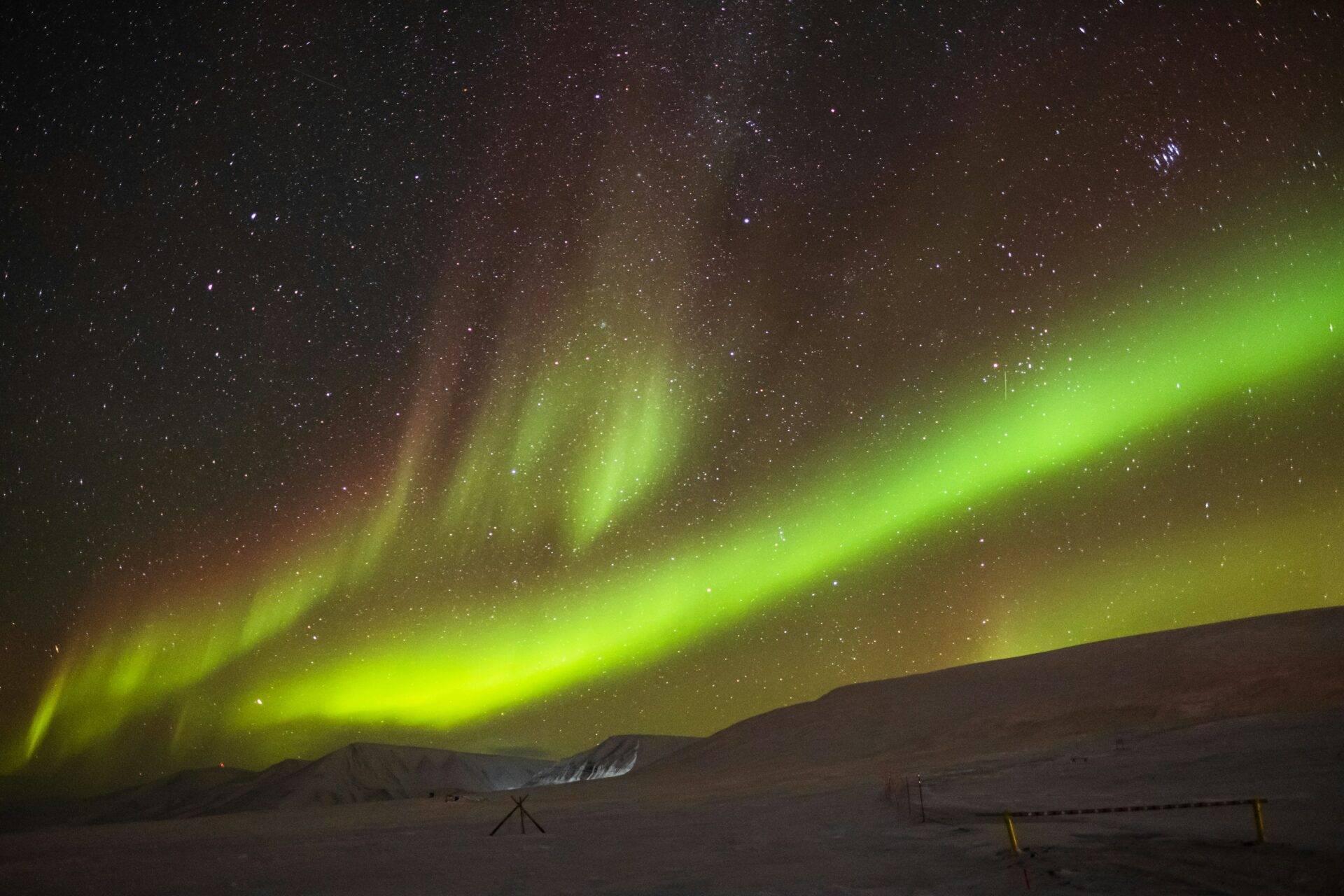 Grüne Nordlichter tanzen über einer verschneiten arktischen Landschaft mit vereinzelten Geräten