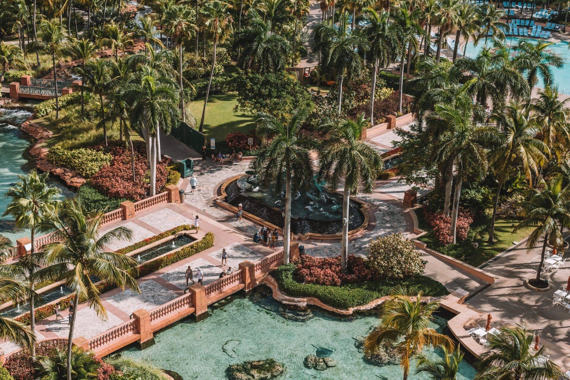 Vista aérea de un resort tropical con piscinas de agua turquesa, terrazas de ladrillo, palmeras y esculturas rodeadas de vegetación