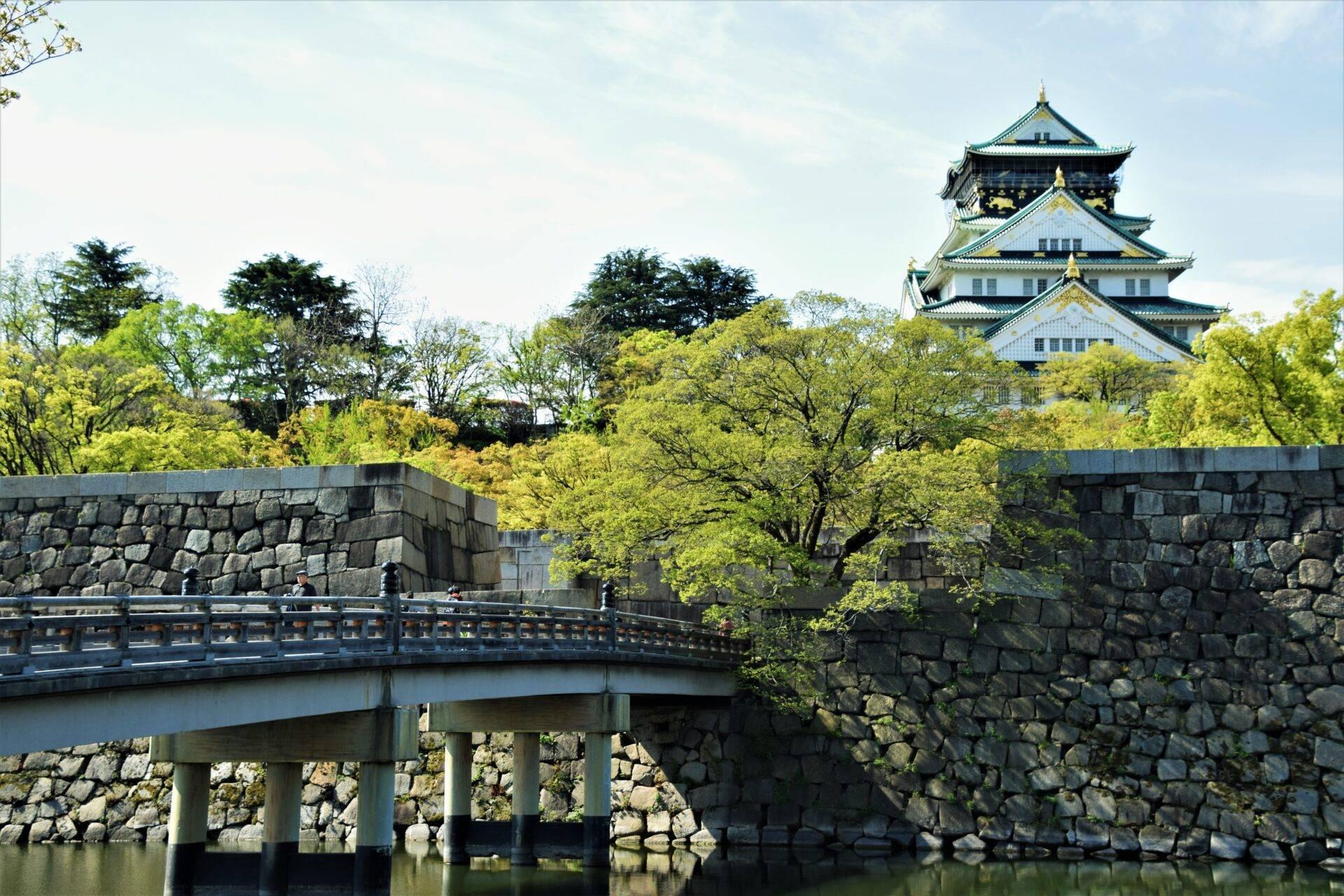 Un château japonais traditionnel aux toits courbes domine un paysage verdoyant traversé par un pont de pierre