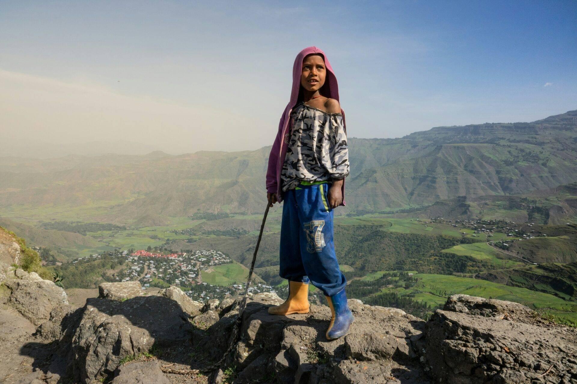 Ein junges Mädchen in traditioneller Tracht posiert auf felsigen Höhen eines grünen, bergigen Tals