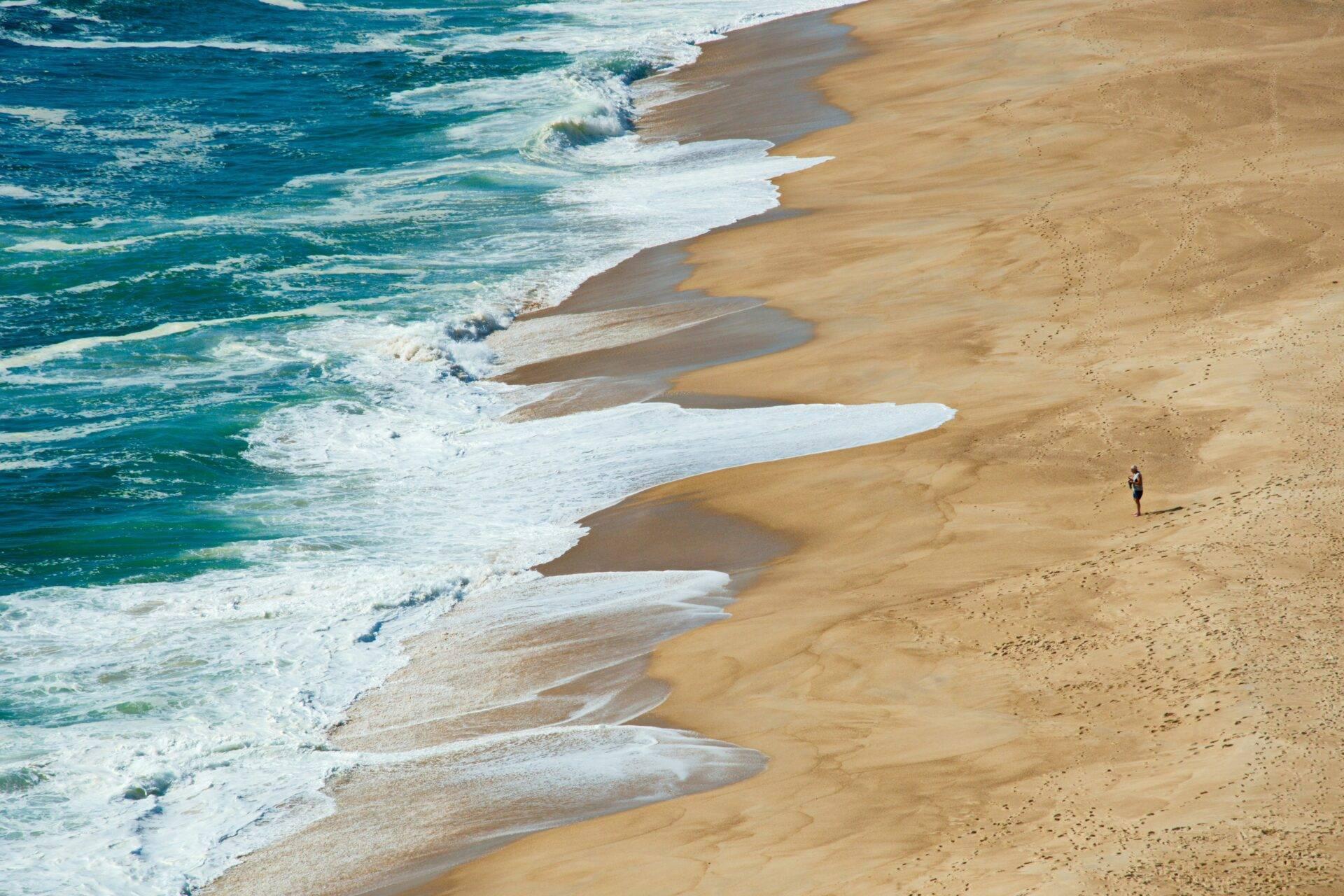 Vista aerea di una spiaggia di sabbia dorata con onde frangenti e un passante solitario in Portogallo