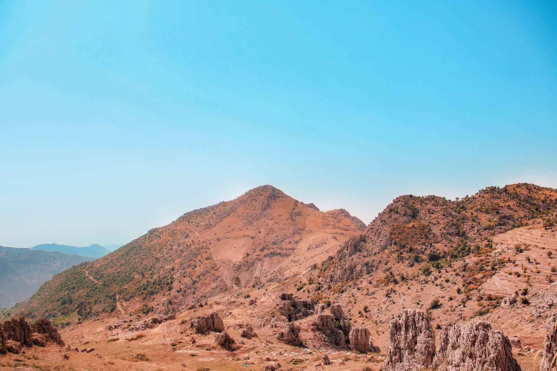 Paysage montagneux aride avec formations rocheuses, végétation clairsemée et chaînes de montagnes en arrière-plan sous ciel dégagé