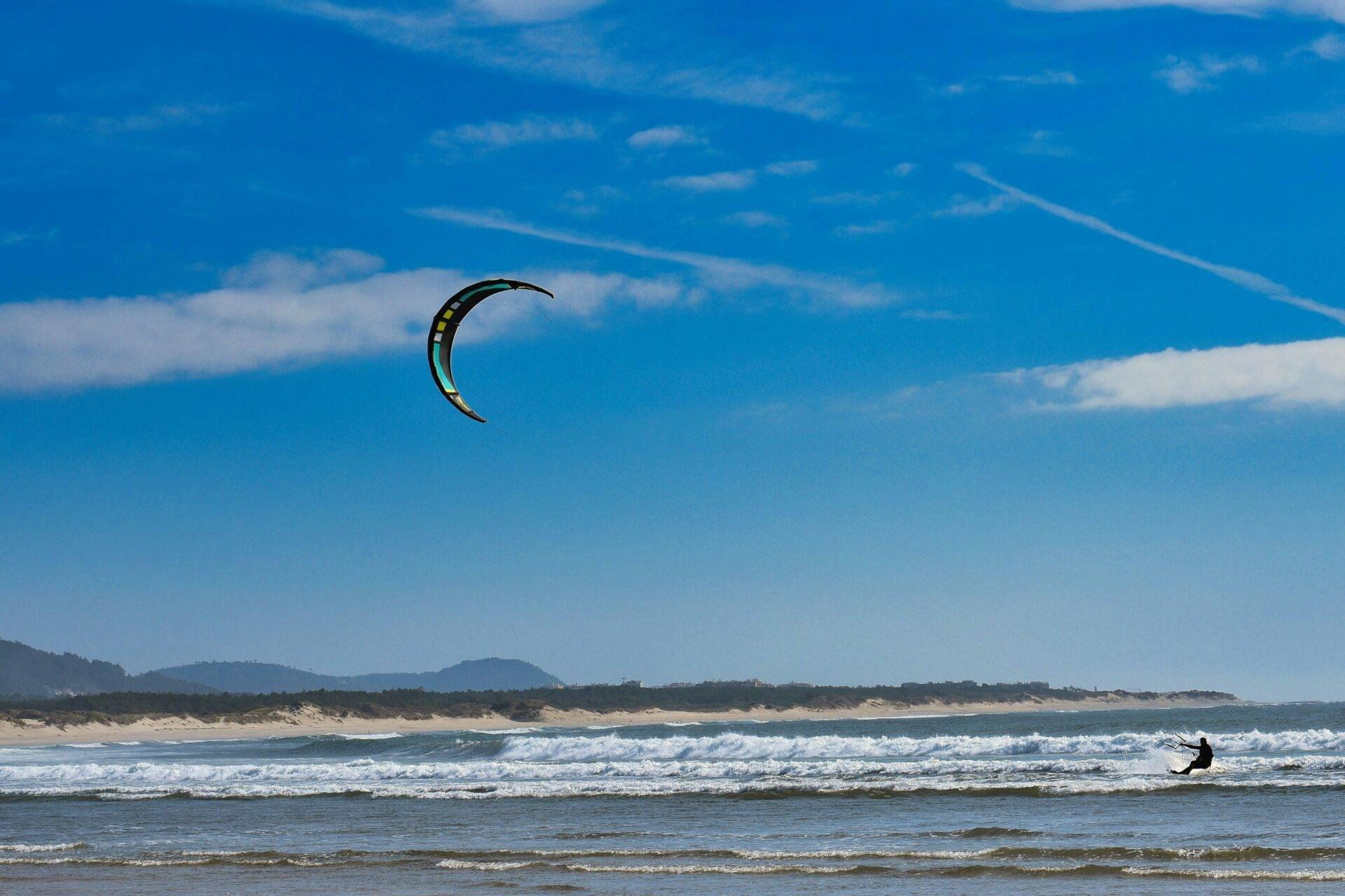 Un kitesurfista in azione su una spiaggia di sabbia fine con onde, sotto un cielo azzurro e montagne sullo sfondo