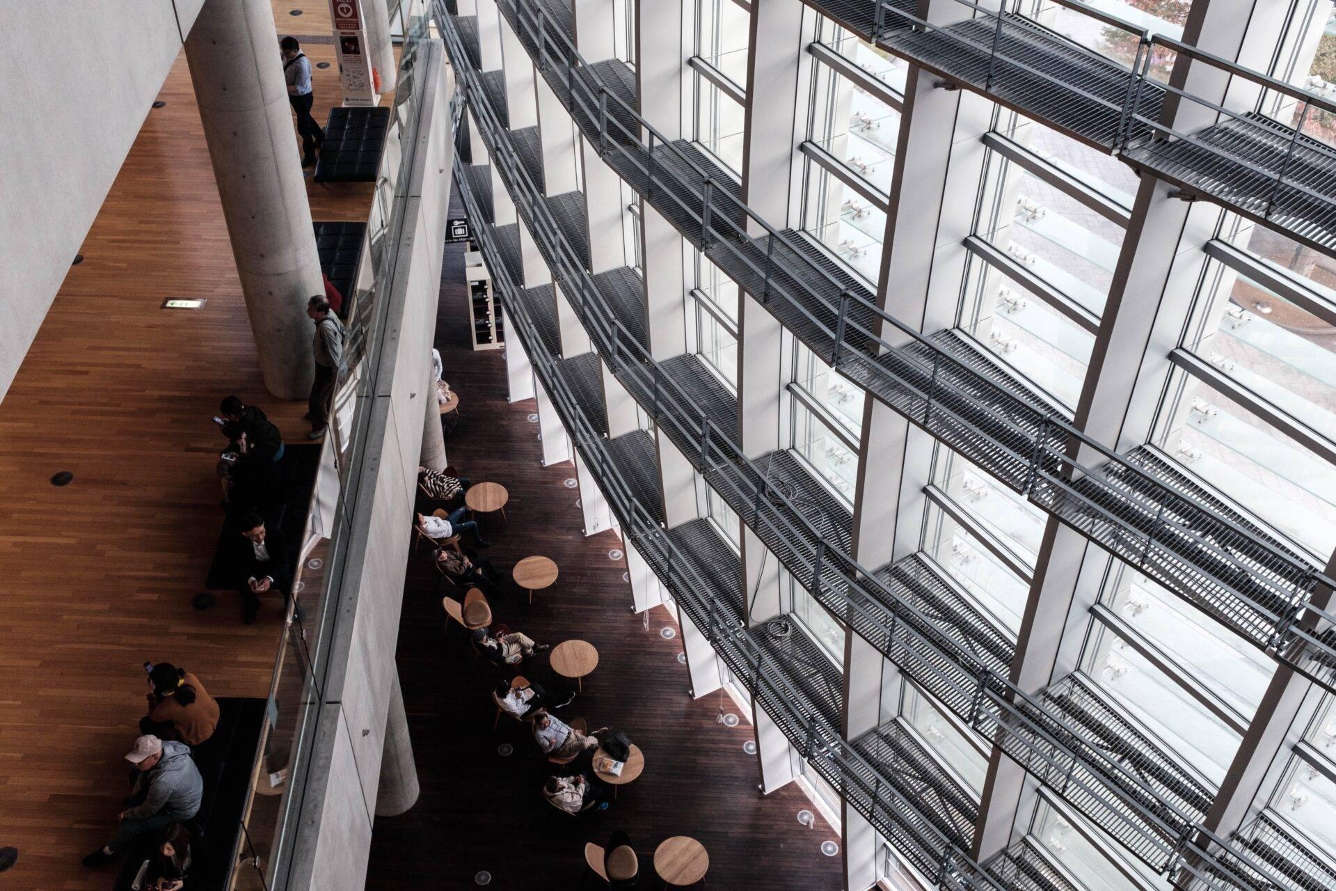 Vue aérienne d’un atrium moderne avec baies vitrées, où des visiteurs sont assis à des tables rondes le long d’une promenade