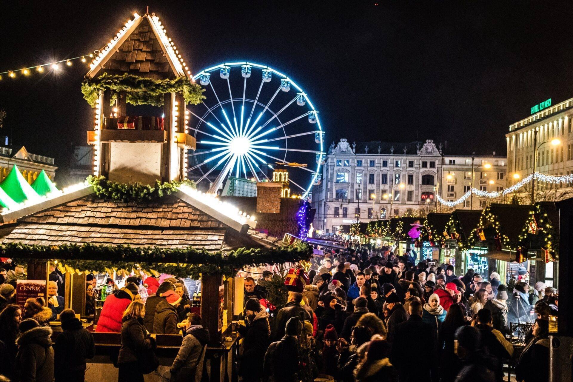 Un marché de Noël illuminé la nuit avec une grande roue bleue, des chalets décorés et une foule festive