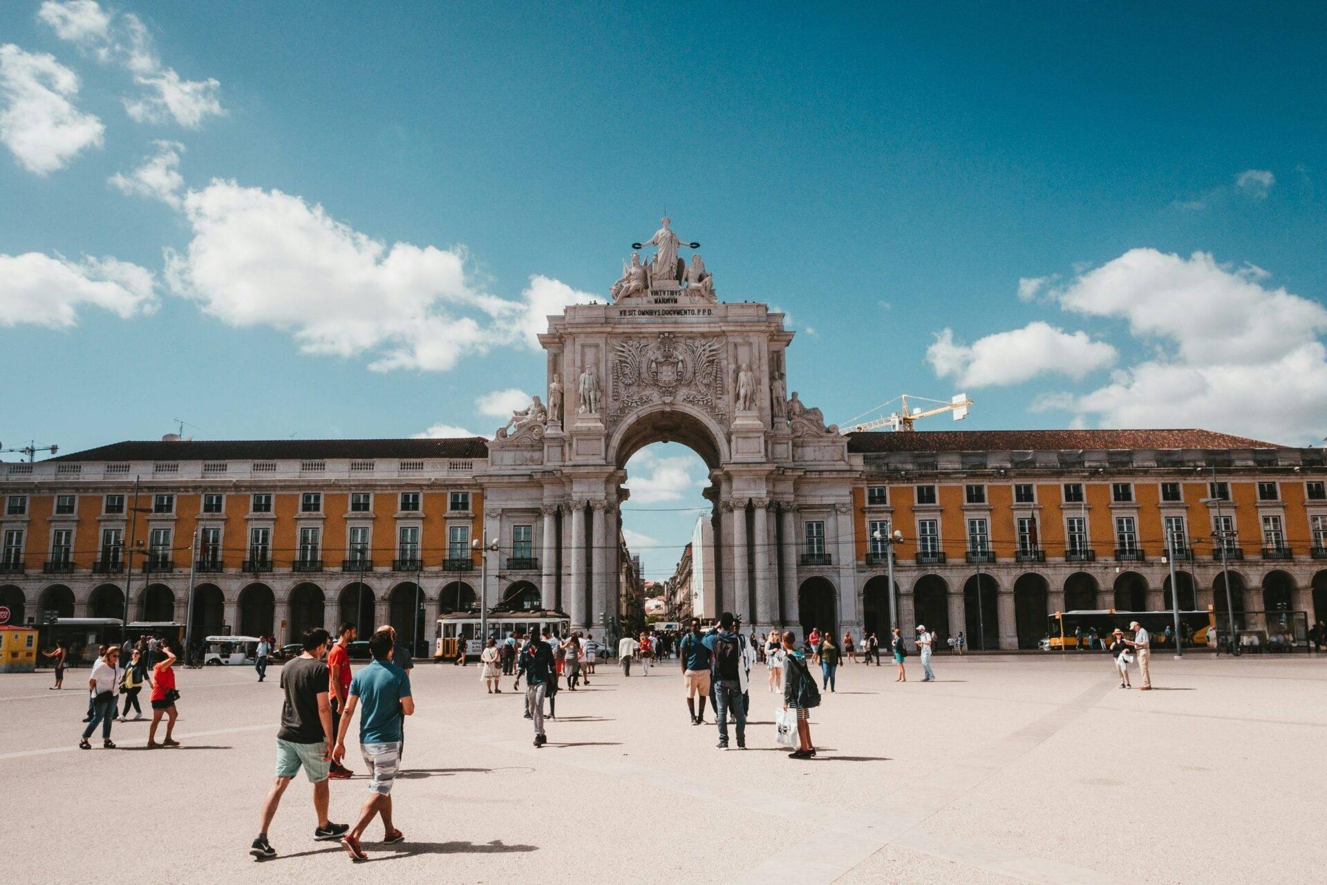 Una grande piazza soleggiata con un monumentale arco trionfale in pietra, bordato da edifici ocra con portici, animata da turisti