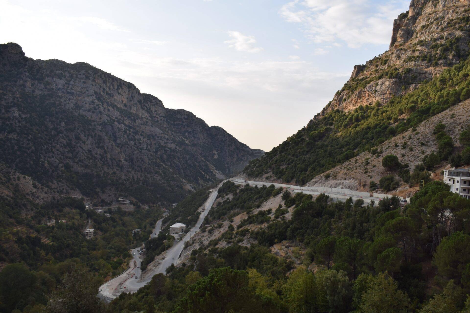 Une route sinueuse traverse une vallée montagneuse avec falaises rocheuses, végétation méditerranéenne et habitations dispersées