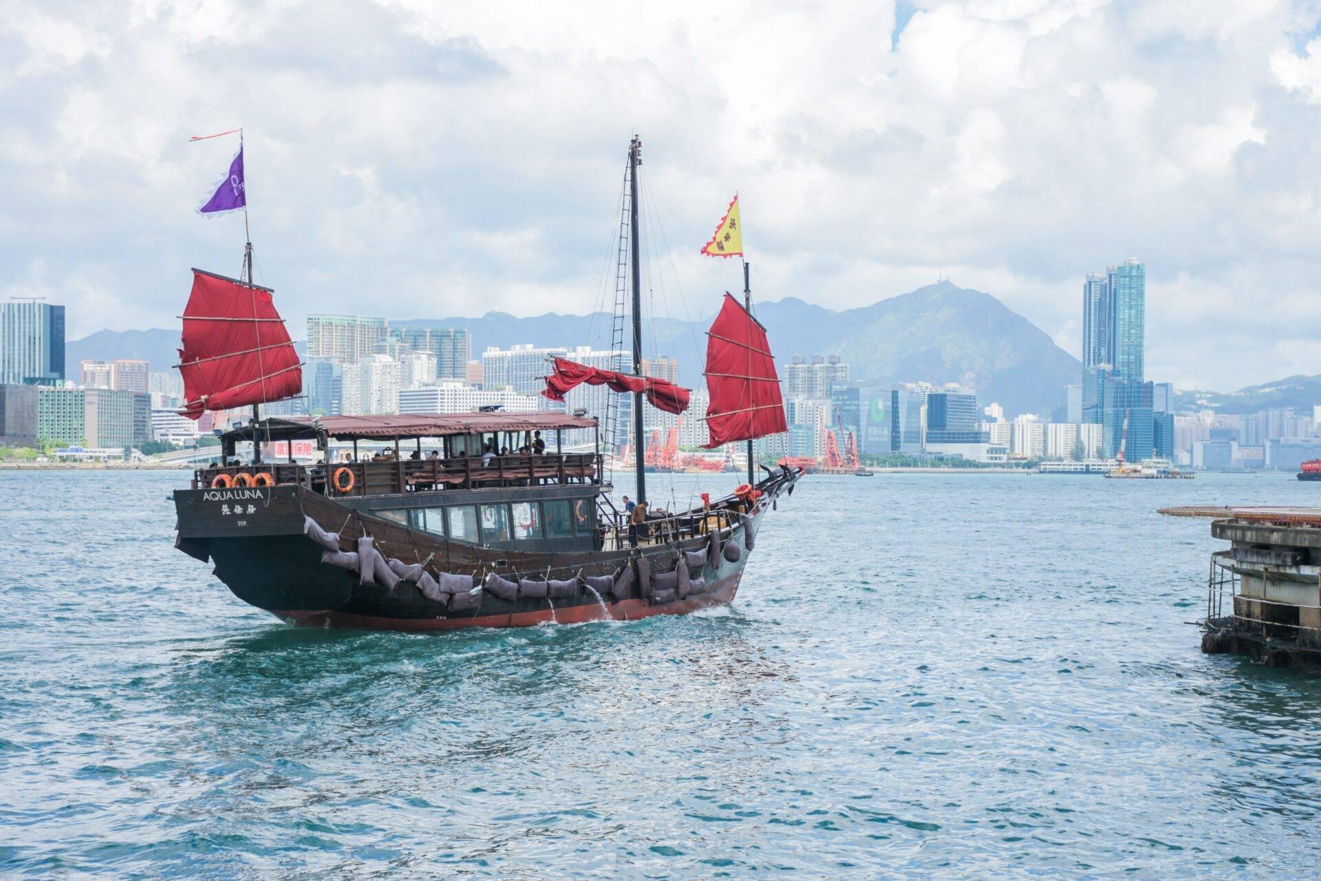 Jonque traditionnelle aux voiles rouges naviguant dans un port urbain avec gratte-ciel et montagnes en arrière-plan