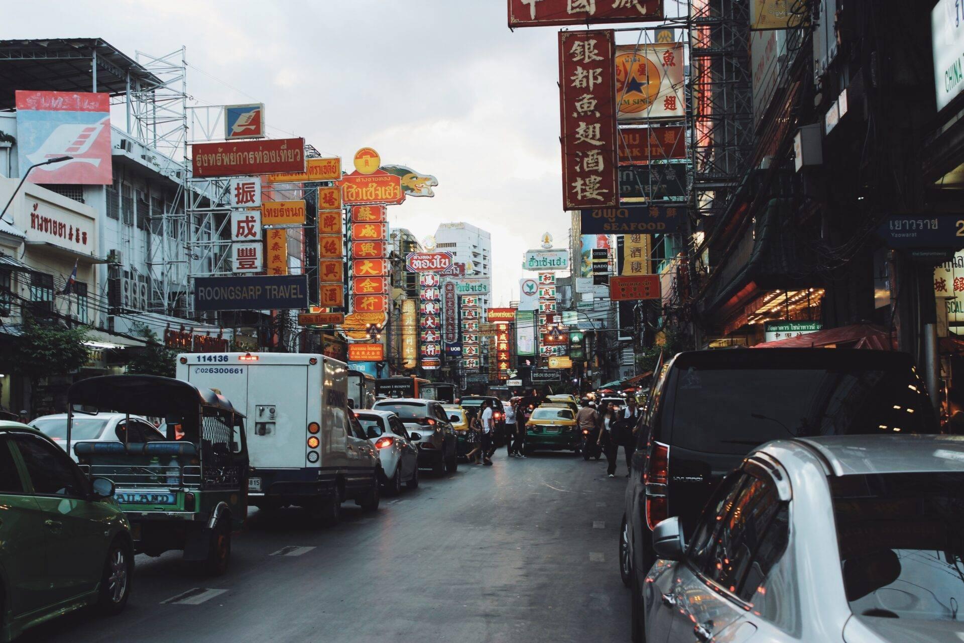 Une rue commerçante animée de Bangkok avec enseignes chinoises colorées, voitures et piétons sous un ciel couvert