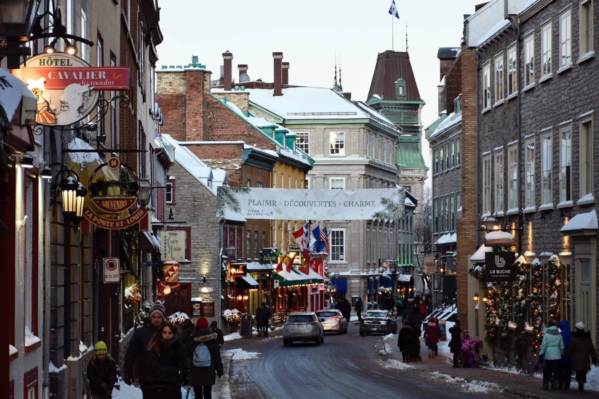 Une rue historique enneigée bordée de bâtiments en pierre avec boutiques décorées, animée par des piétons en période festive