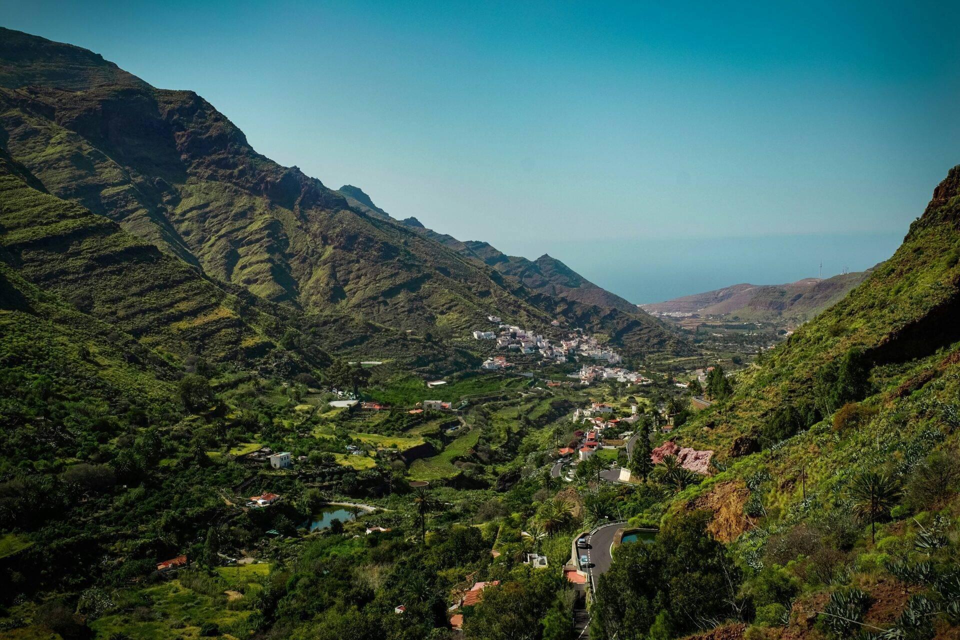 Vista aerea di una valle verdeggiante con villaggi bianchi, strada tortuosa e montagne scoscese sotto un cielo sereno