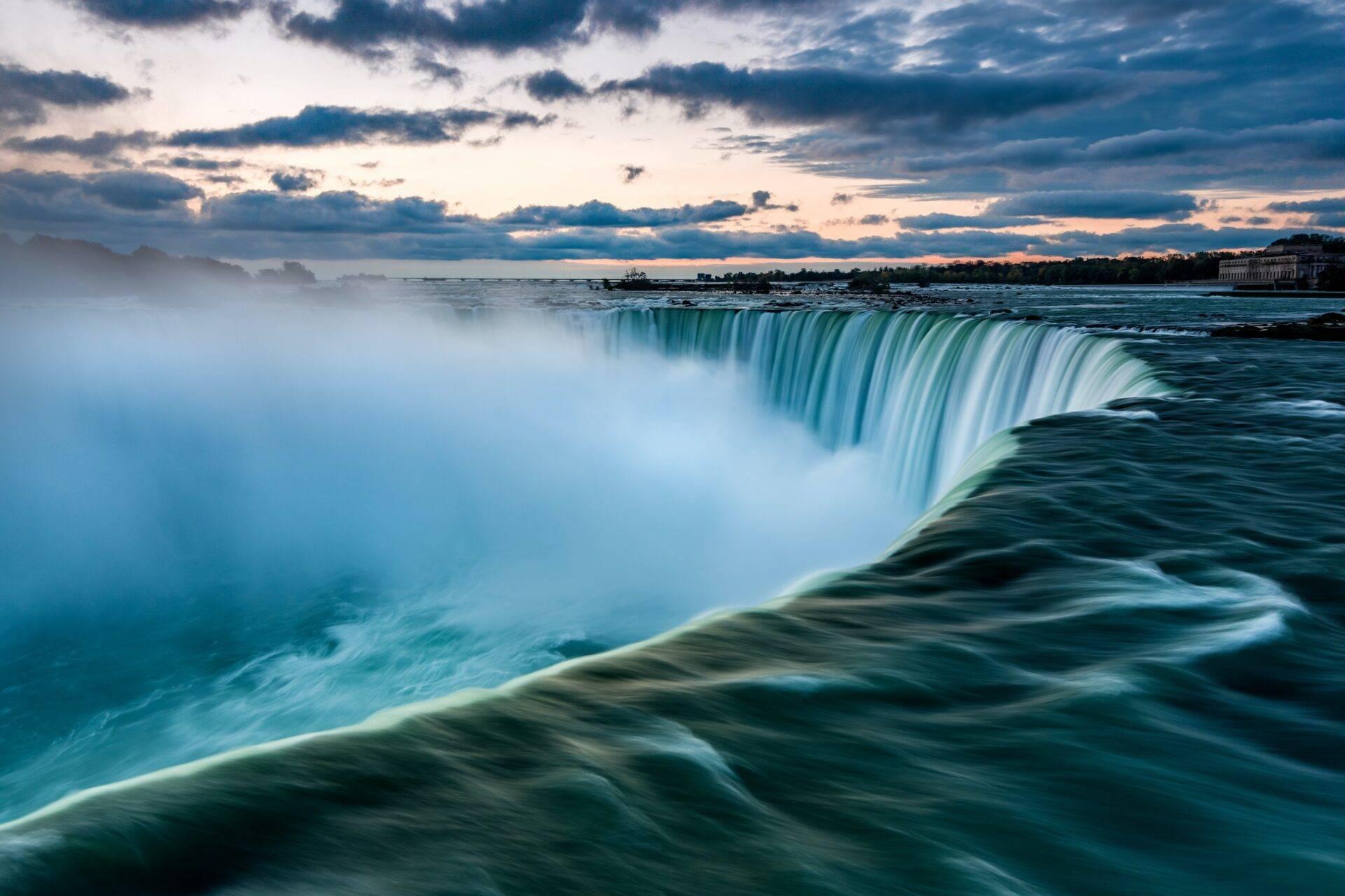 Une majestueuse cascade d’eau turquoise se déverse dans une immense chute d’eau sous un ciel nuageux au coucher de soleil