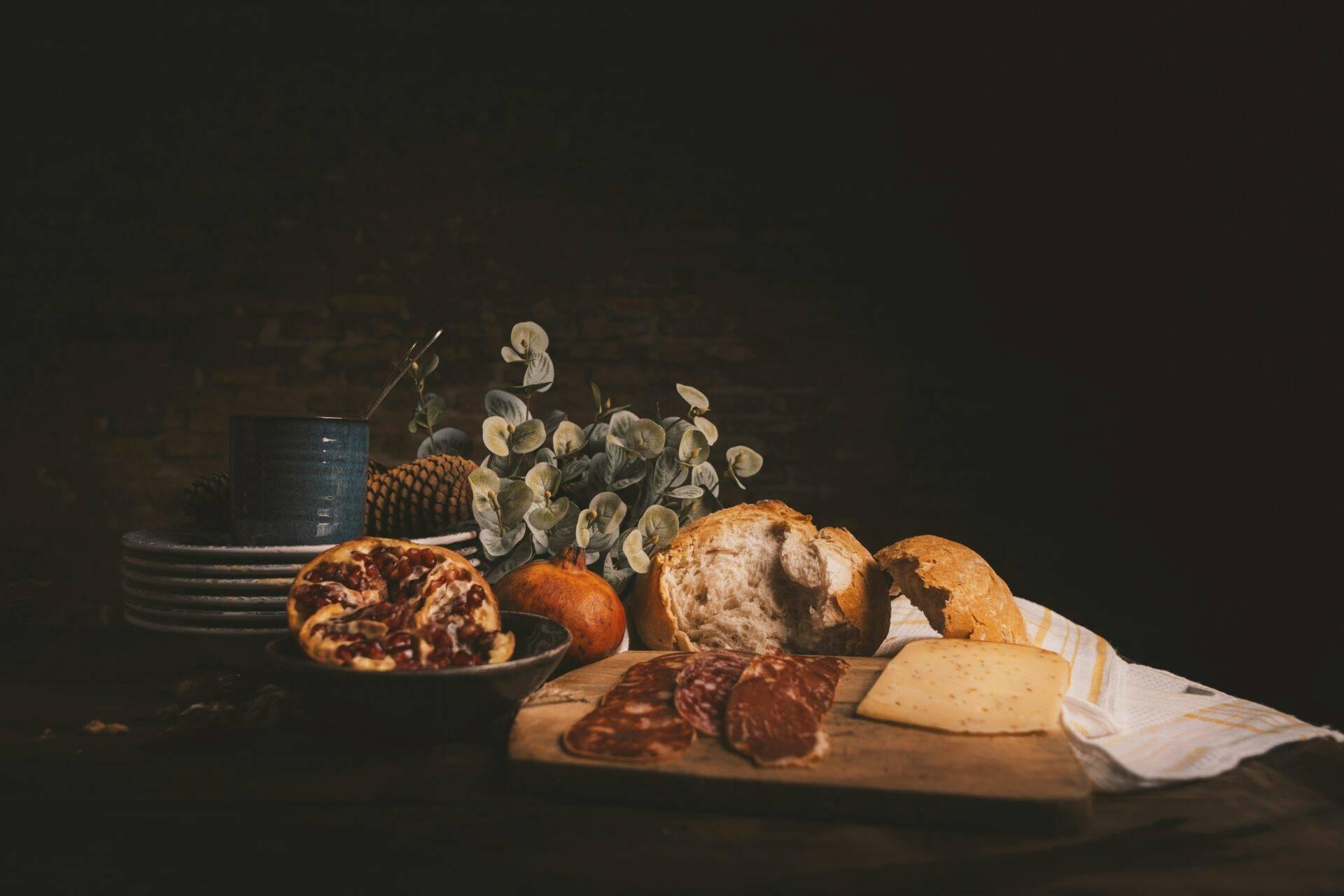 Composizione culinaria con pane, affettati, formaggio, frutta e fiori essiccati su sfondo scuro