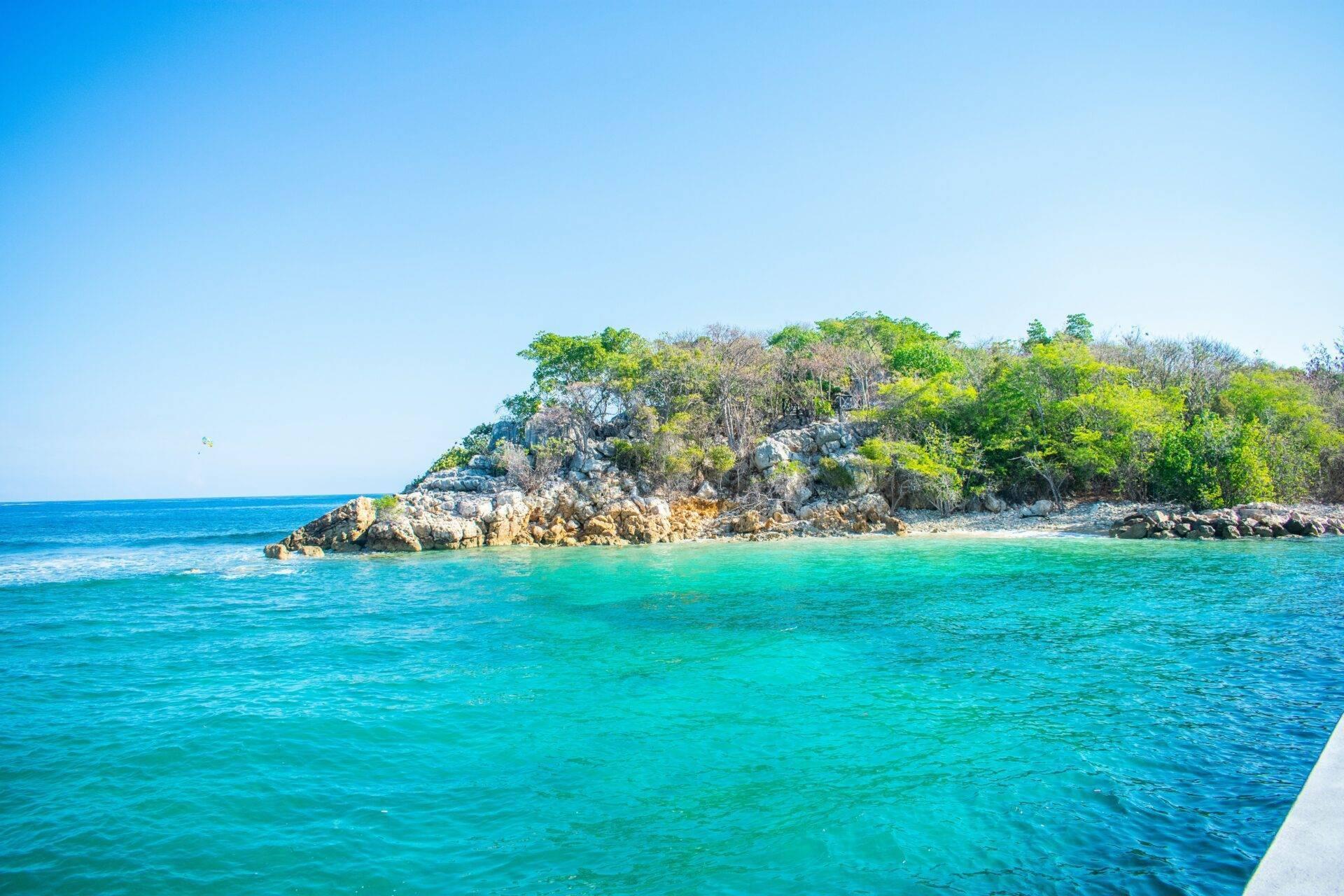 Una piccola isola rocciosa ricoperta di vegetazione tropicale emerge dalle acque turchesi cristalline sotto un cielo sereno