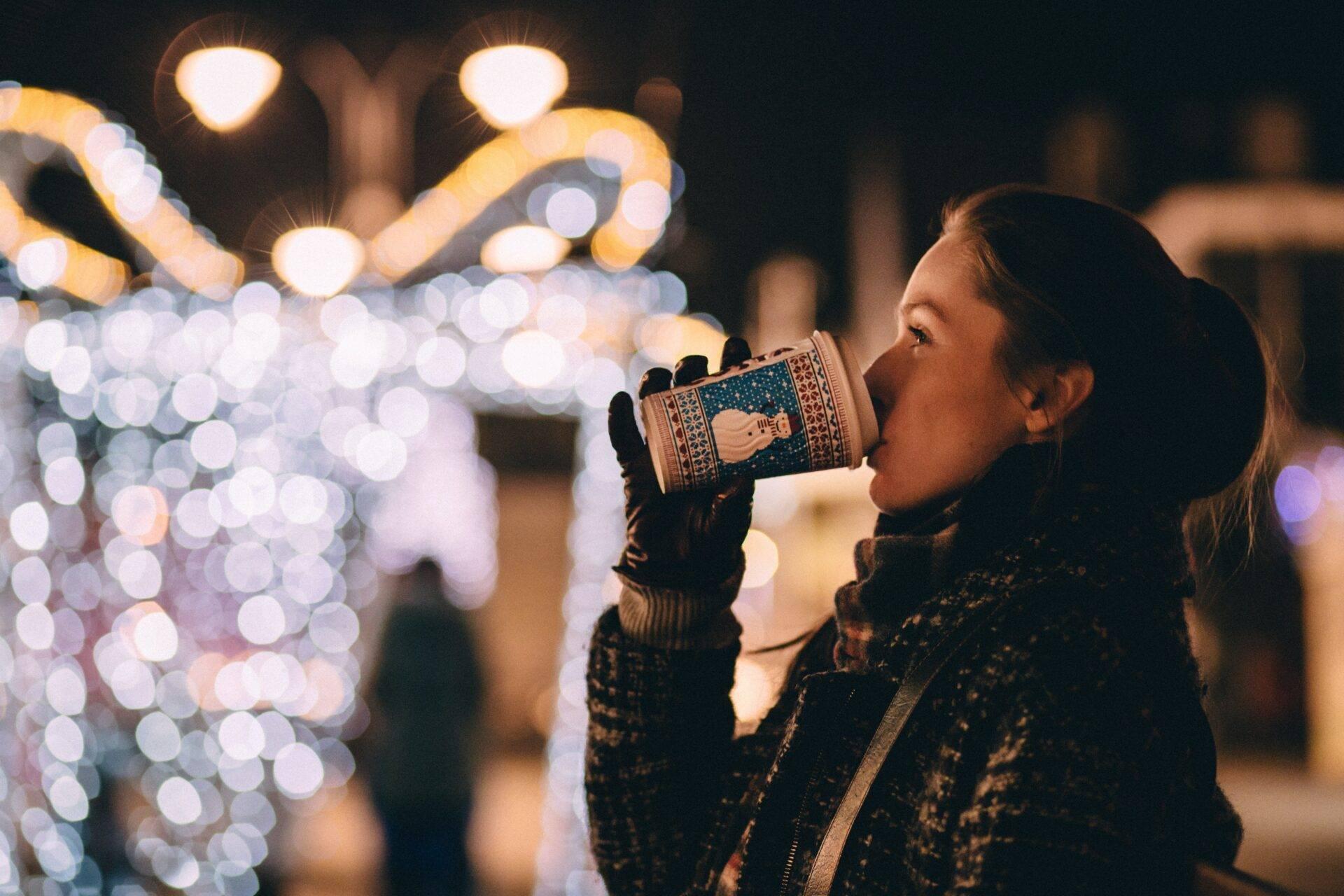 Une jeune femme boit une boisson chaude devant des illuminations de Noël scintillantes en ville la nuit