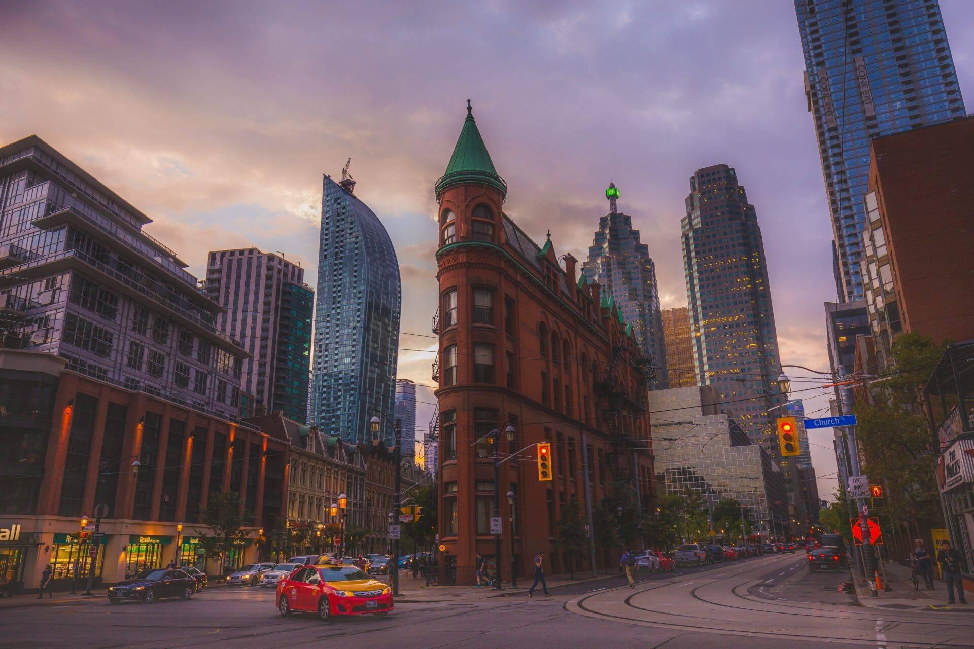 Un bâtiment historique en brique rouge avec tours vertes dominant une intersection urbaine au crépuscule, entouré de gratte-ciel modernes