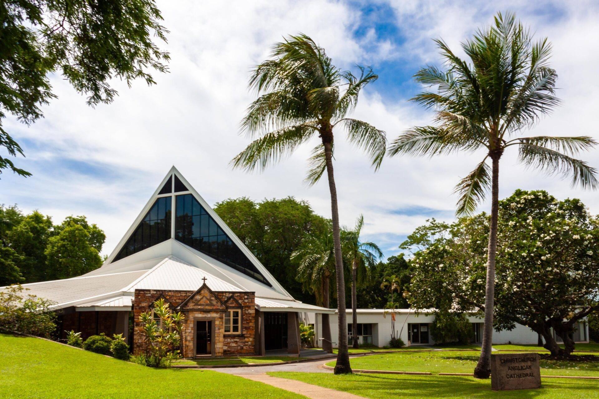 Una iglesia moderna con tejado blanco en punta y cruz, rodeada de palmeras y vegetación bajo un cielo azul