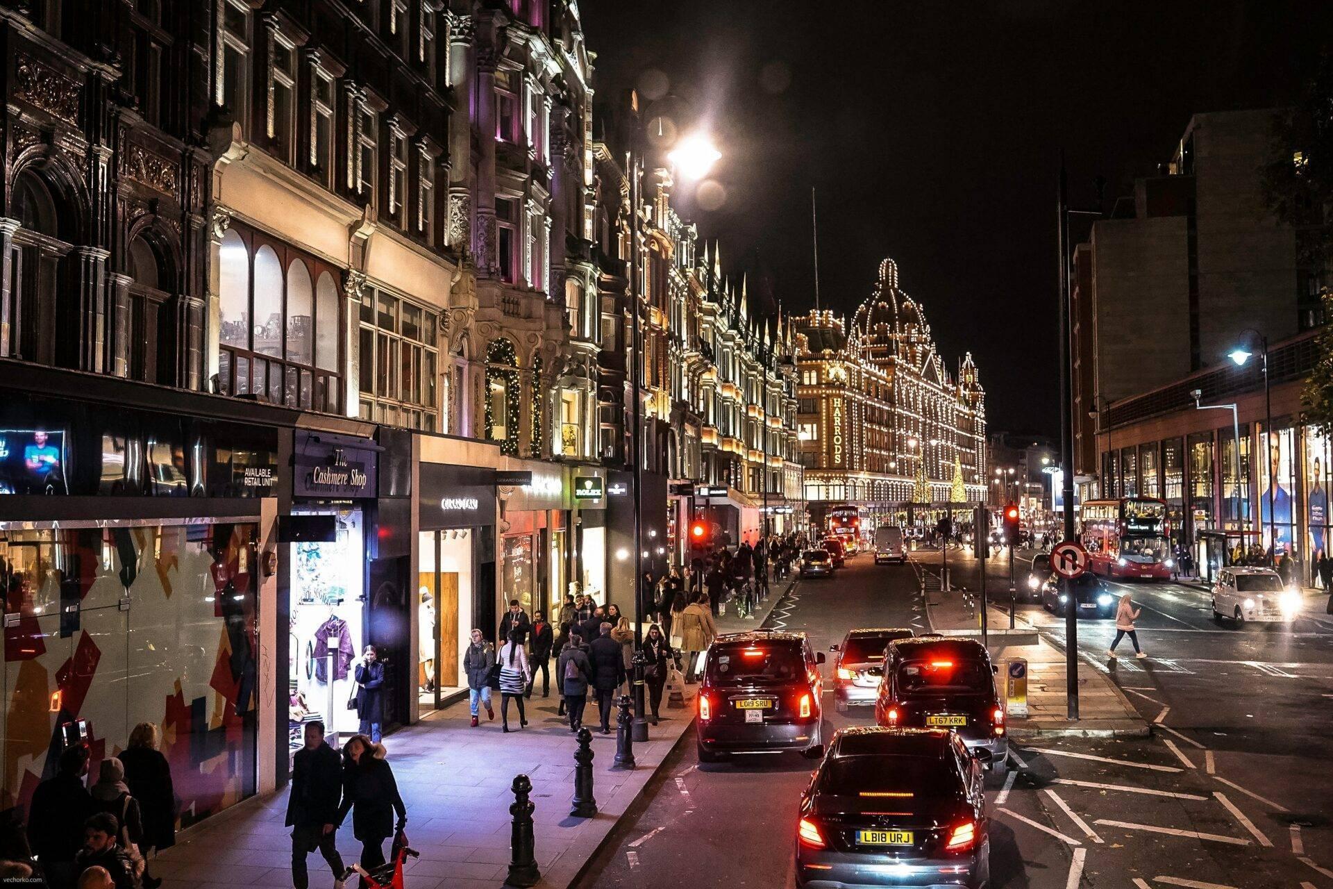 Une rue commerciale animée la nuit, illuminée pour les fêtes, avec façades victoriennes, boutiques et taxis londoniens