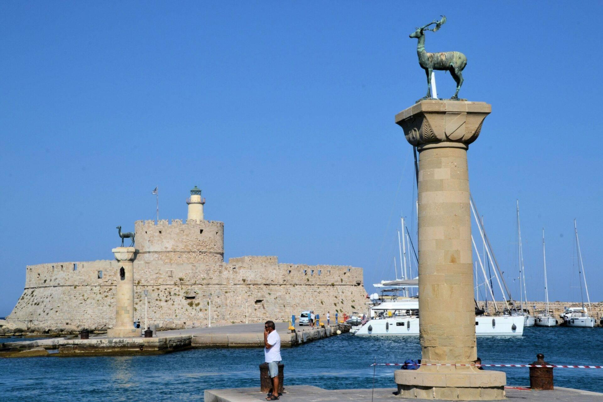 Un port méditerranéen avec colonne surmontée d’une statue de cerf, forteresse en pierre et voiliers amarrés
