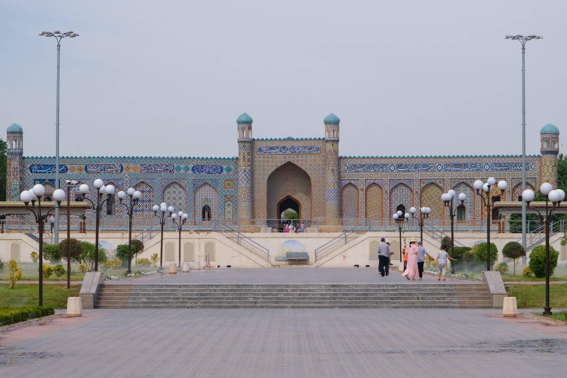 Grande mosquée avec façade ornementale bleue et dorée, minaret central, cour pavée et visiteurs en vêtements légers