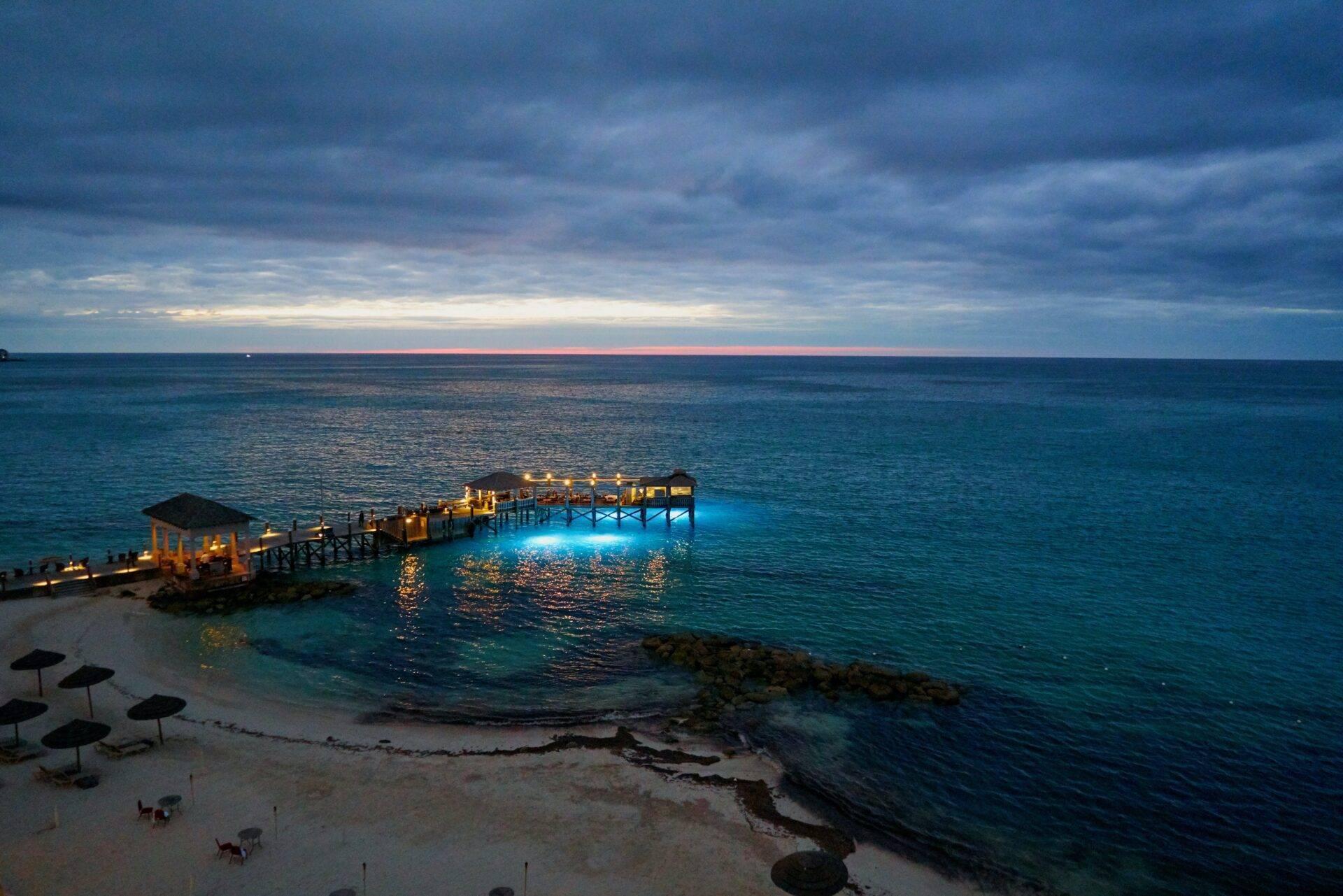 Un embarcadero iluminado adentrándose en un mar turquesa al atardecer, con playa y sombrillas