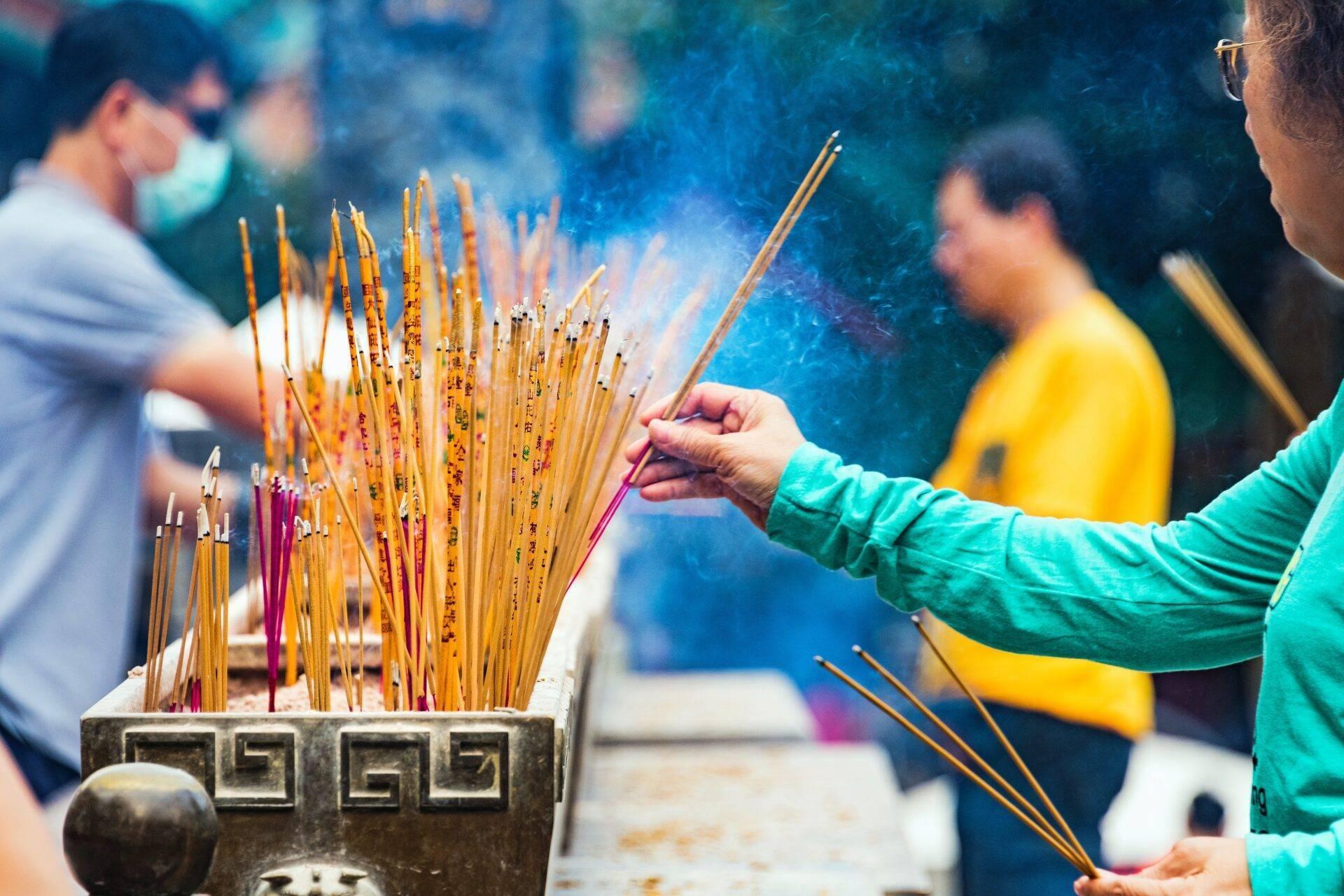 Une personne en veste verte saisit un bâton d’encens dans un brûle-encens orné, en présence de visiteurs dans un temple asiatique
