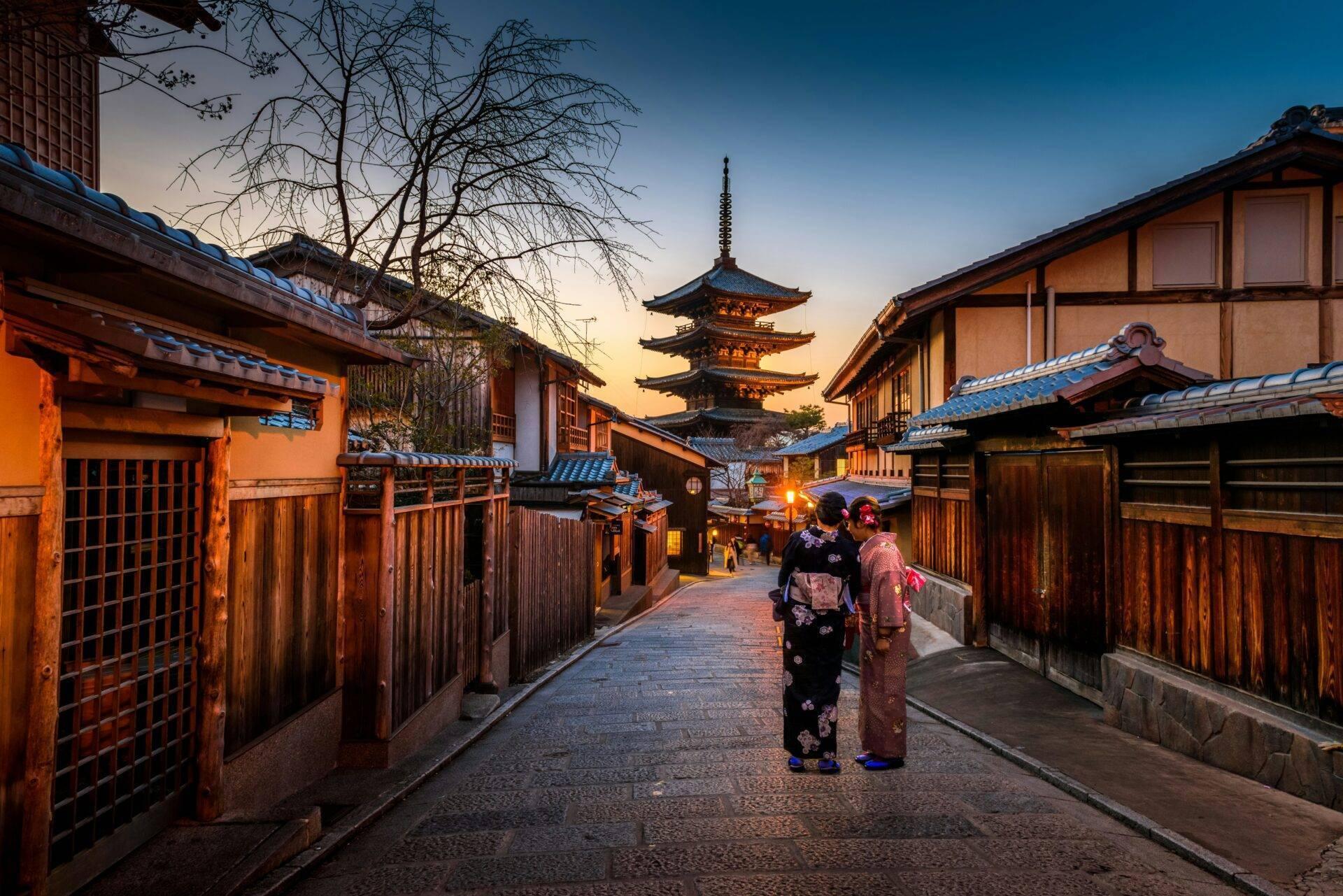 Rue historique pavée au crépuscule avec pagode illuminée, maisons traditionnelles en bois et deux femmes en kimono