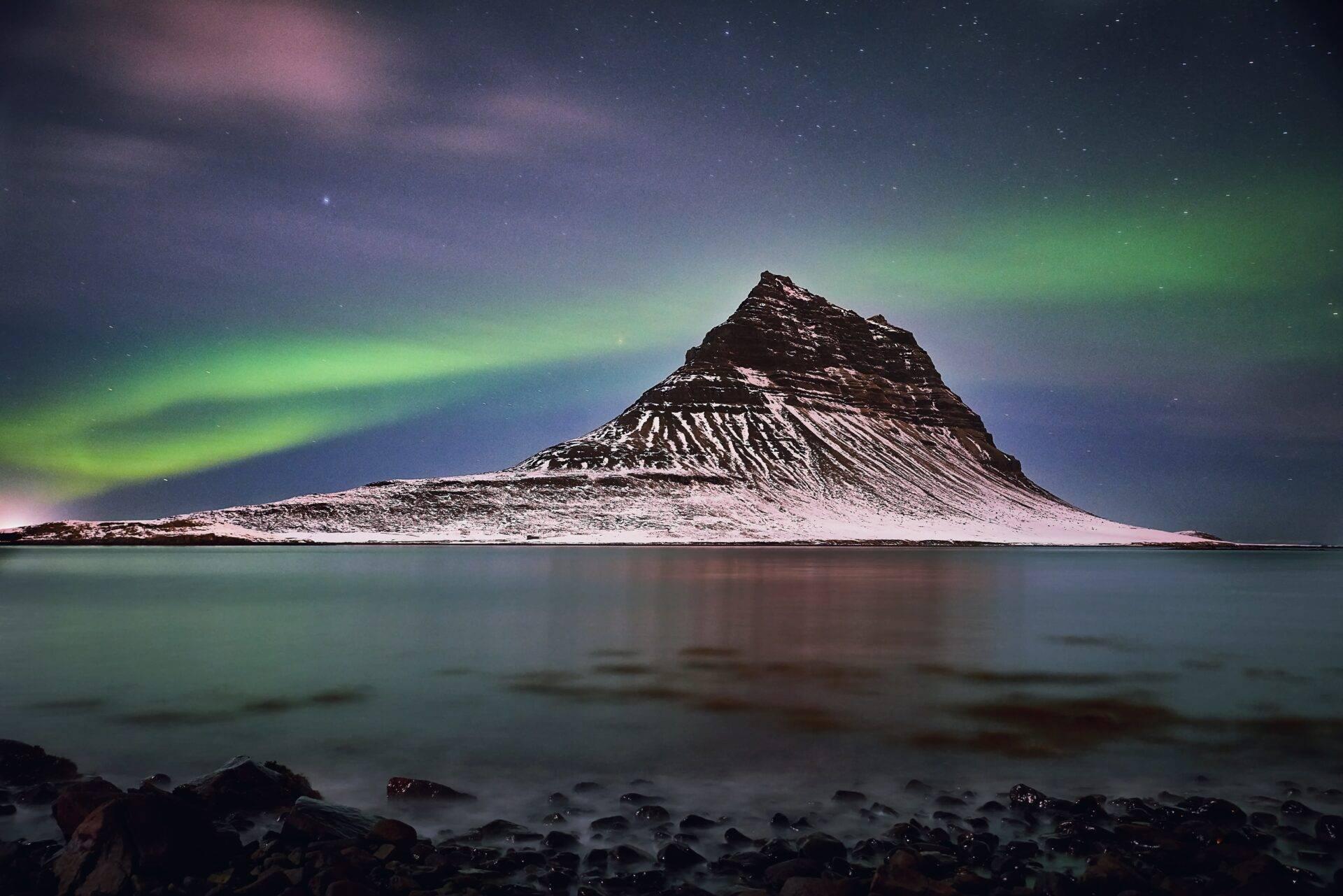 Una montagna innevata si riflette in una baia glaciale sotto un’aurora boreale verde e un cielo stellato