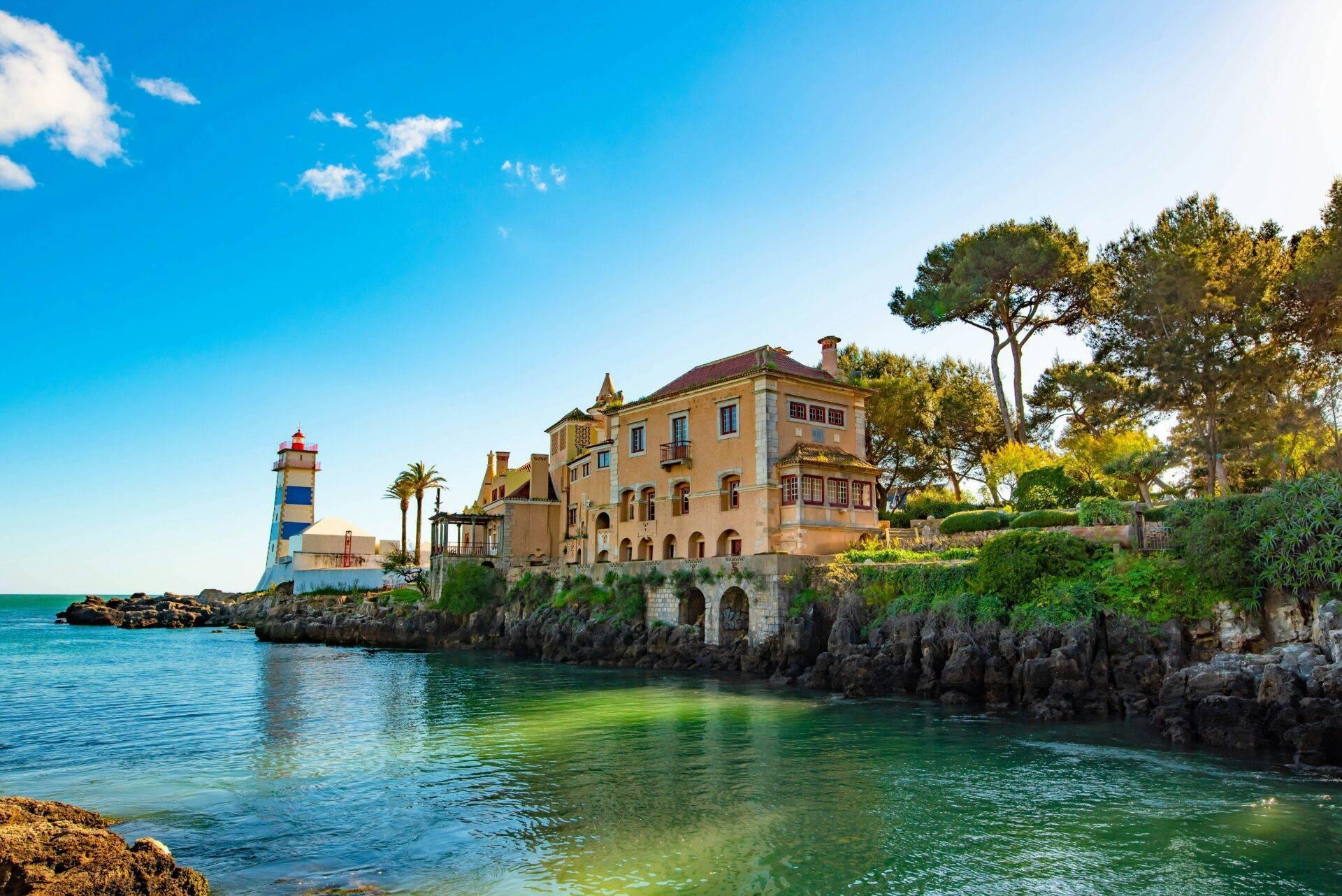 Un palazzo ocra arroccato su rocce costiere con un faro bianco e azzurro che domina una baia turchese mediterranea