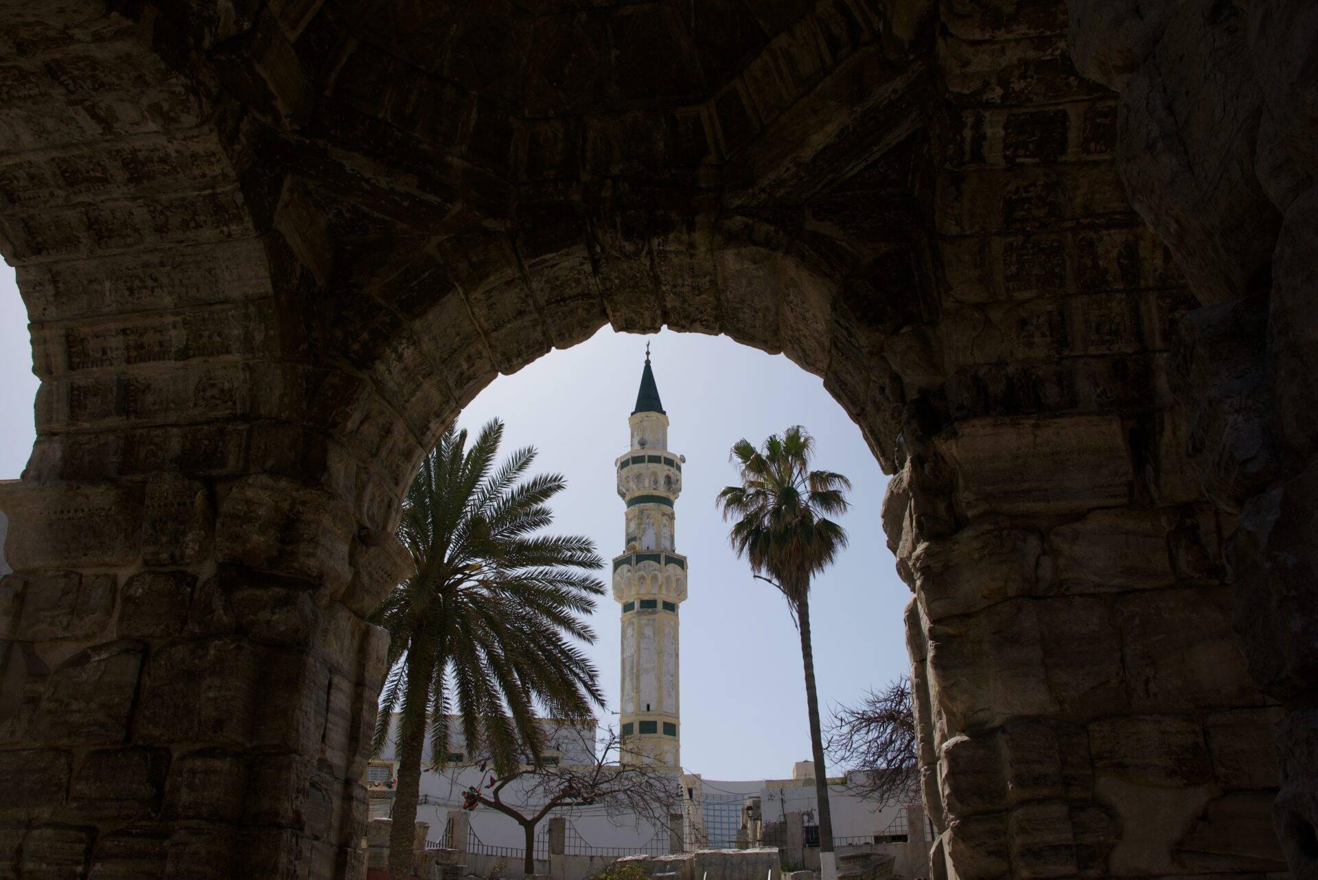 Vue à travers une arche en pierre d’un minaret blanc avec balcons bleus et palmiers