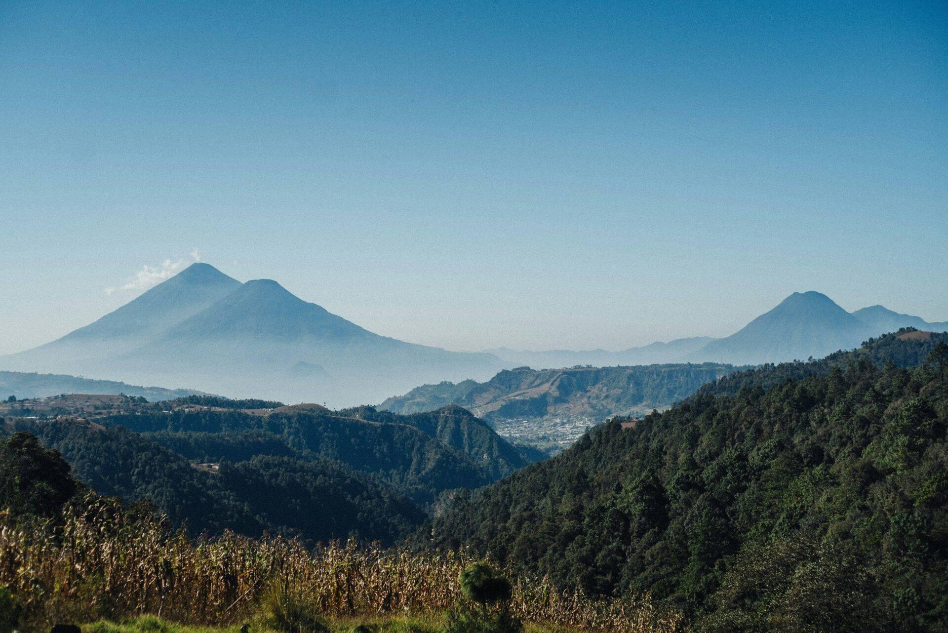 Paysage montagneux du Guatemala avec deux volcans enneigés à l’horizon, forêts de pins et champs agricoles