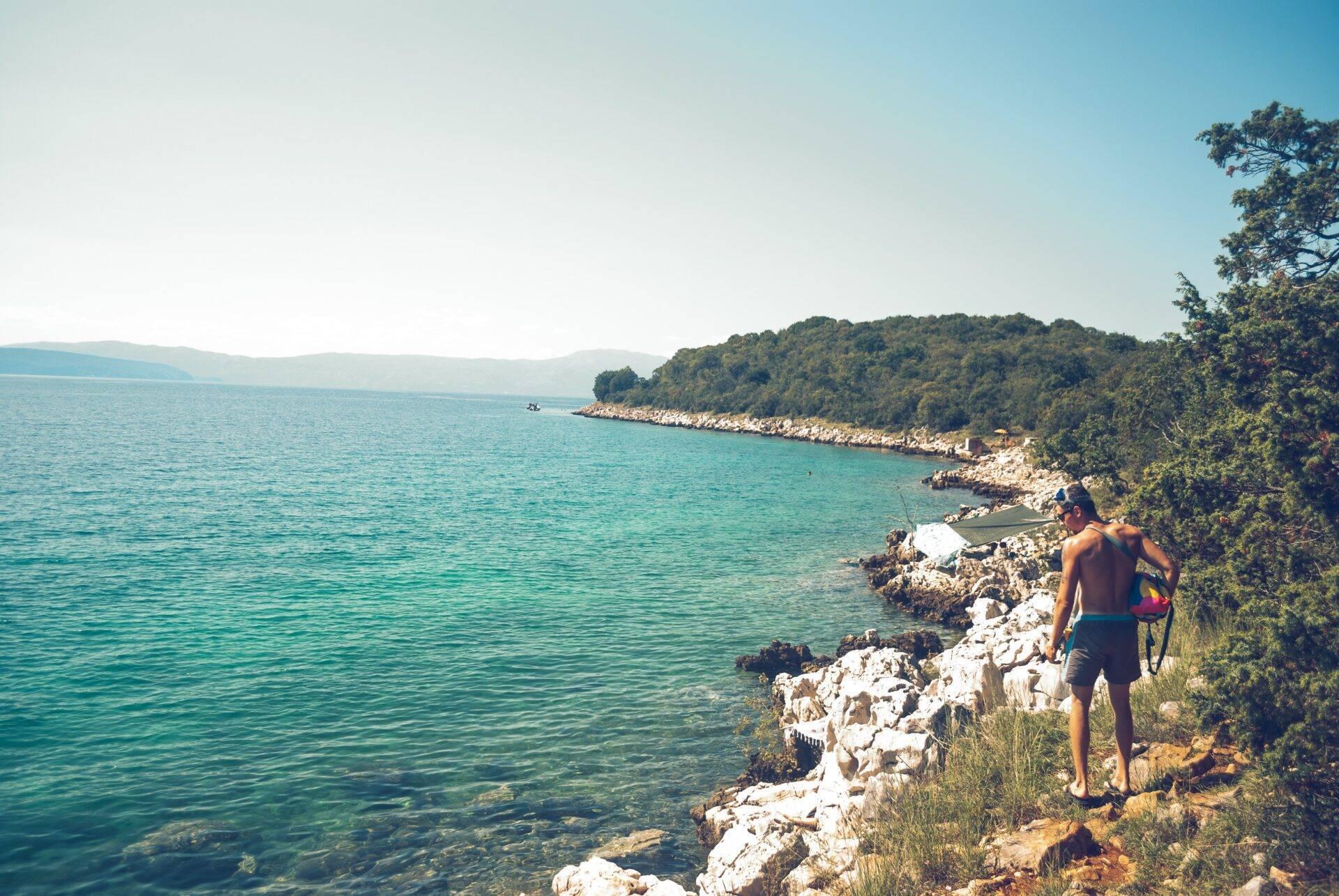 Ein junger Mann in Badehose steht auf Kalksteinfelsen vor einer türkisfarbenen Bucht, umgeben von mediterranem Wald