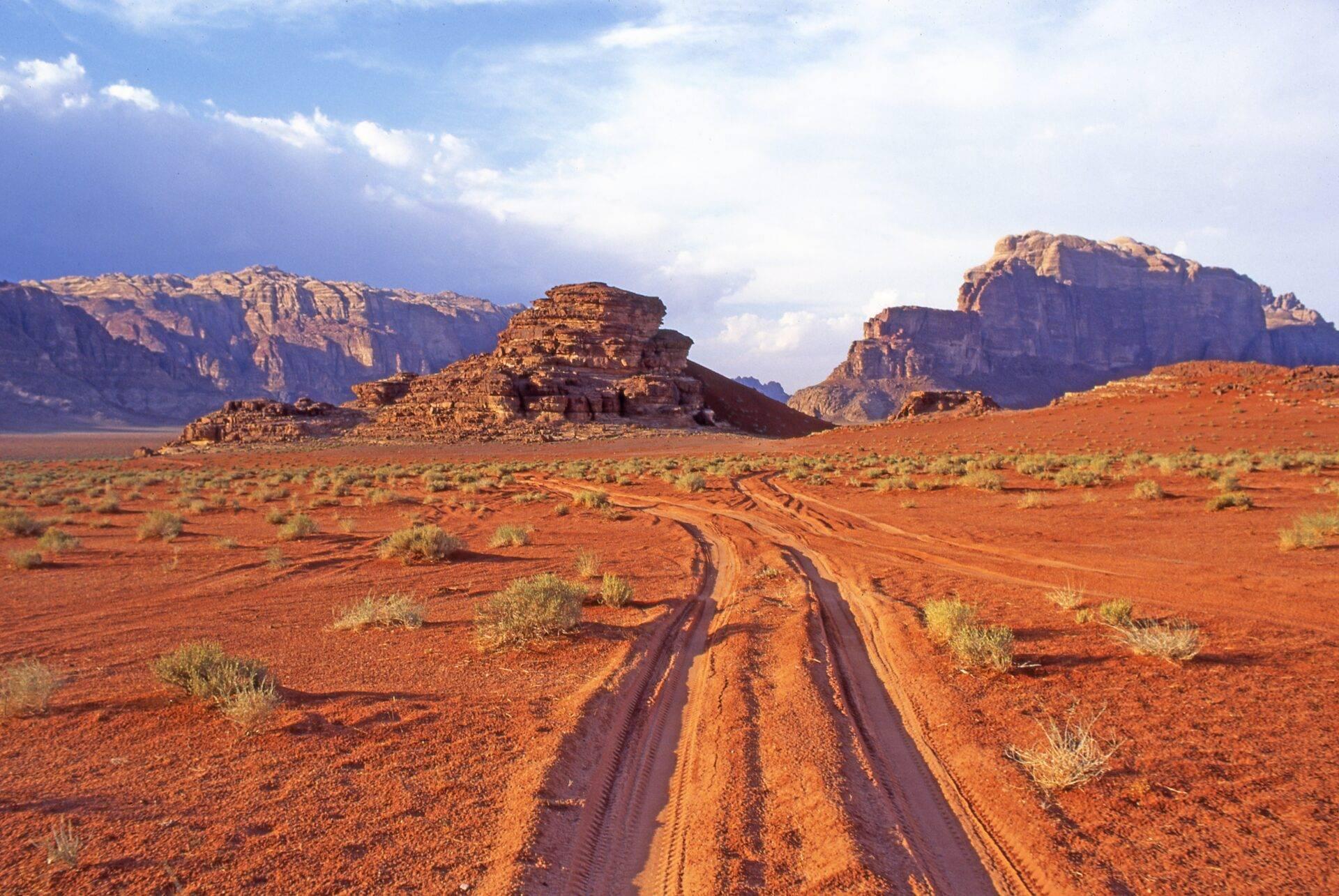 Strada di terra rossa che attraversa un deserto arido con formazioni rocciose monumentali sotto un cielo blu nuvoloso